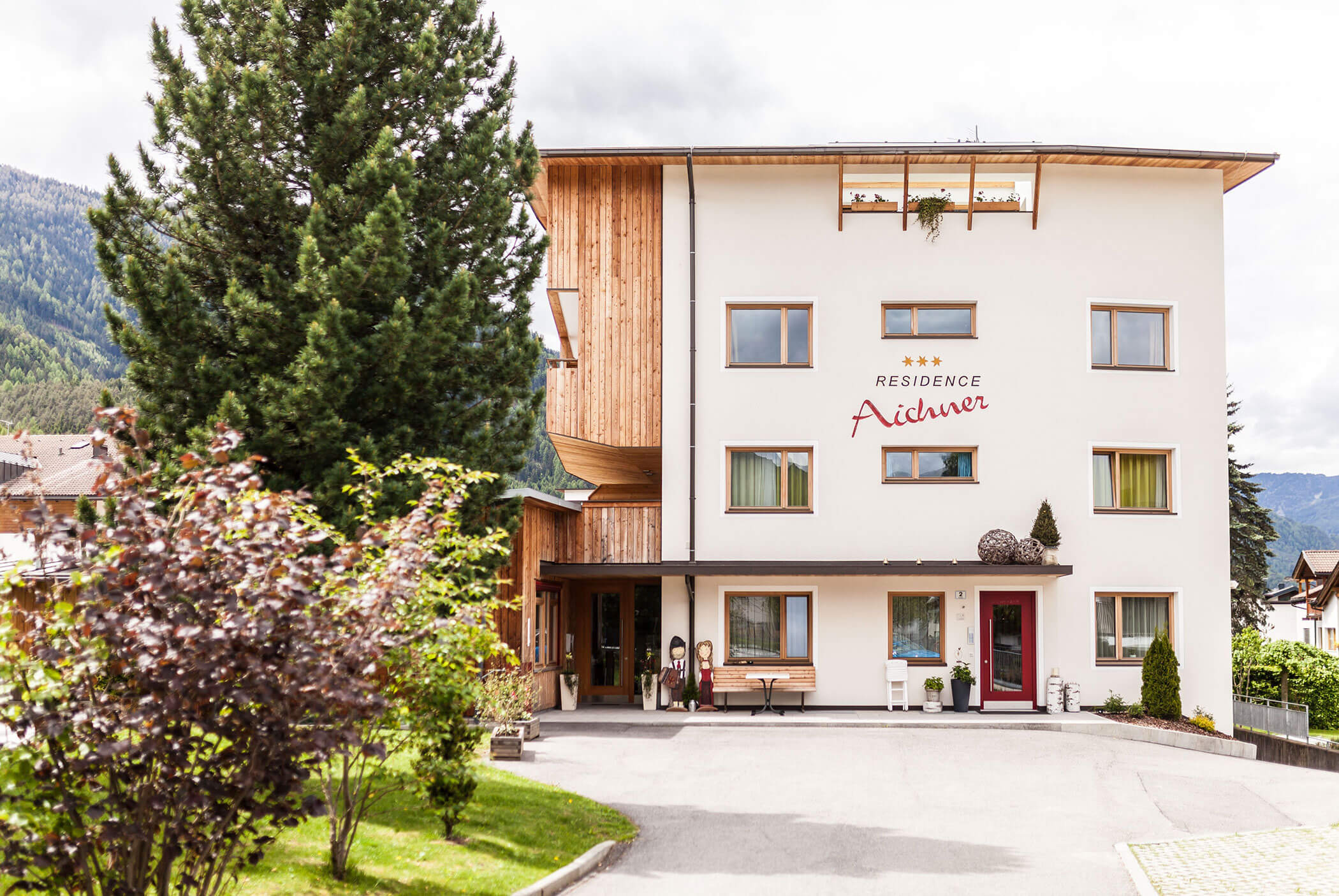 Ein dreistöckiges Hotel namens „Residence Aiduner“ mit Balkonen, Bäumen und Bergen im Hintergrund. - Residence Aichner