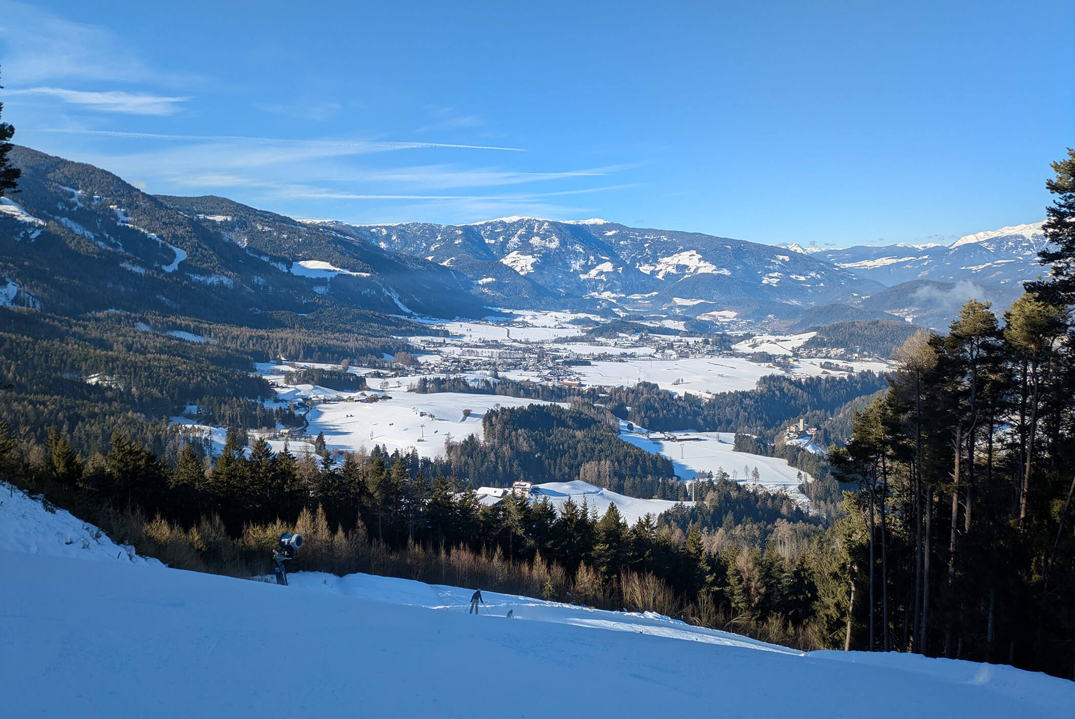 Snowy valley with forests and mountains under a clear blue sky, skiers visible in the foreground. - Residence Aichner