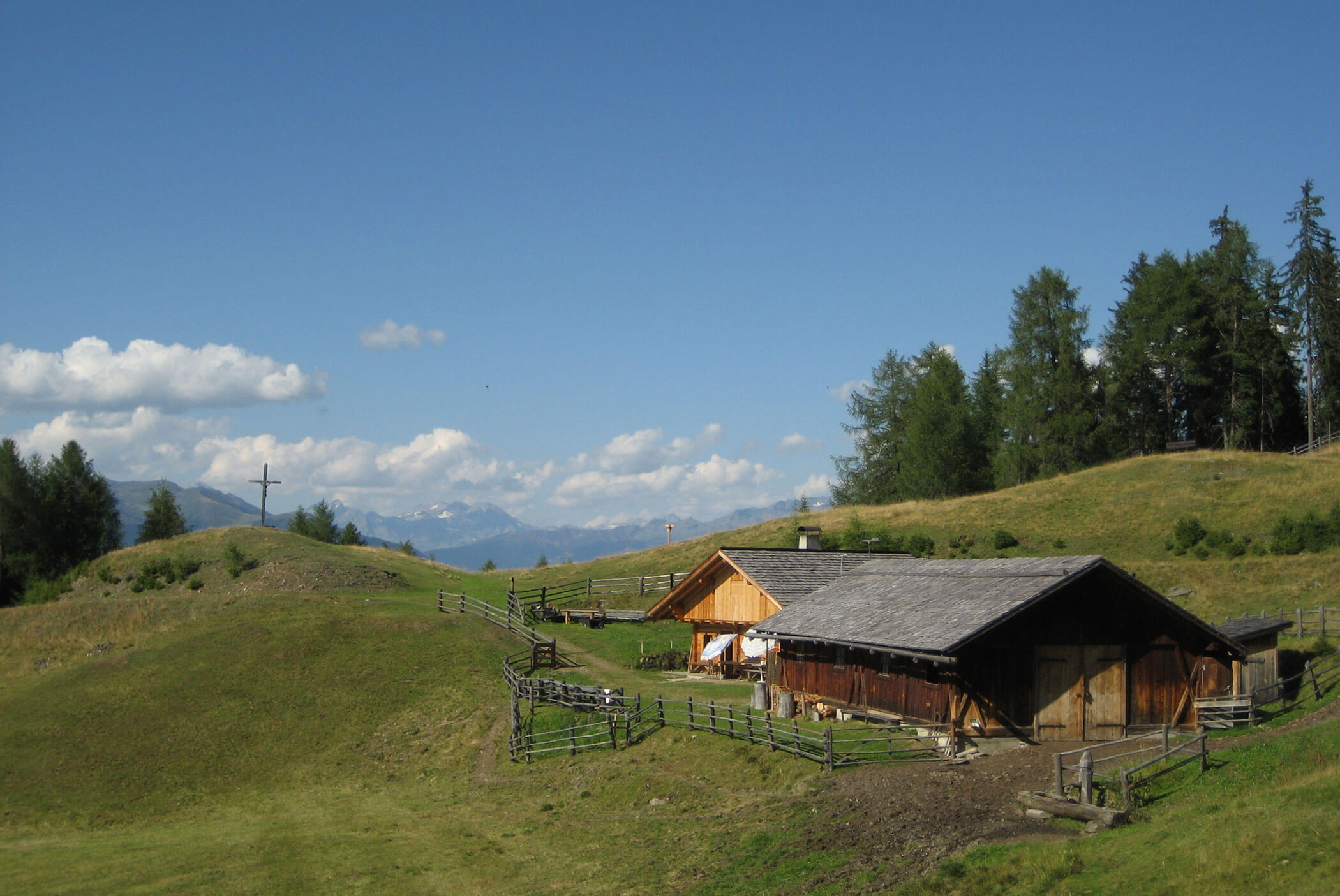 Wooden cabins on a grassy hillside with trees, a cross, and distant mountains under a blue sky. - Residence Aichner