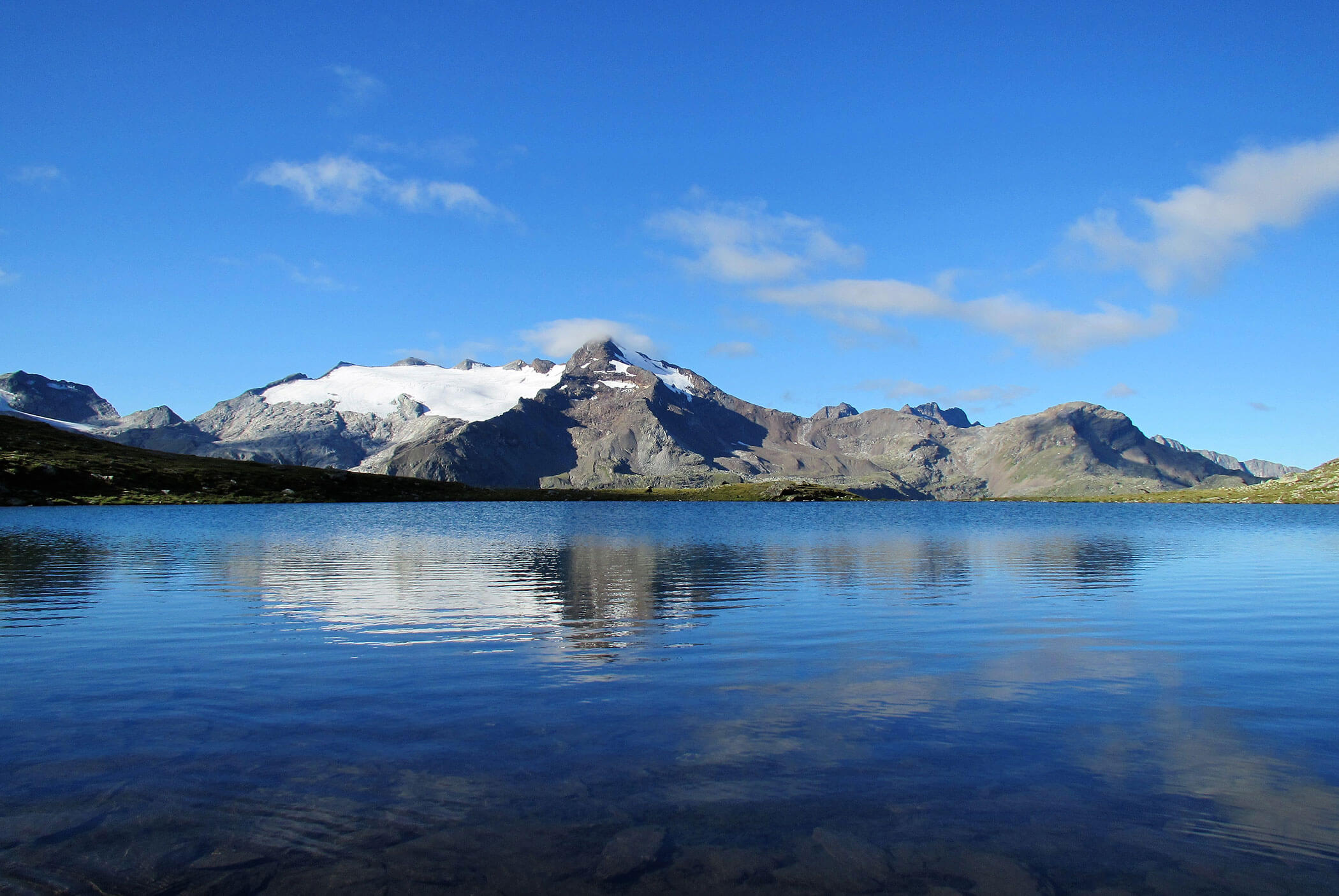Schneebedeckte Berge spiegeln sich in einem klaren, blauen See unter einem strahlenden Himmel mit vereinzelten Wolken. - Residence Aichner