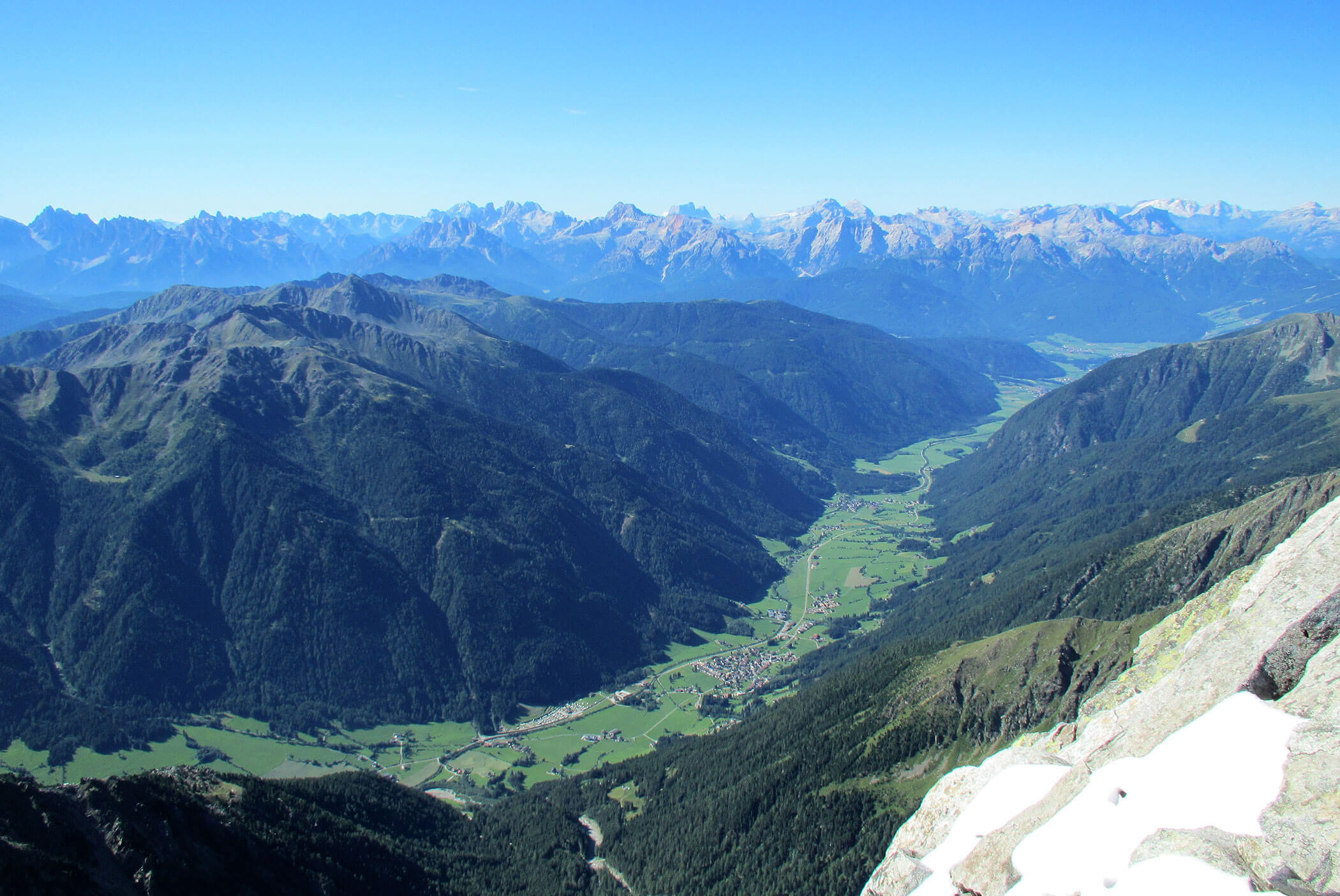 A panoramic view of a mountain valley with green fields, forests, and distant peaks under a clear blue sky. - Residence Aichner