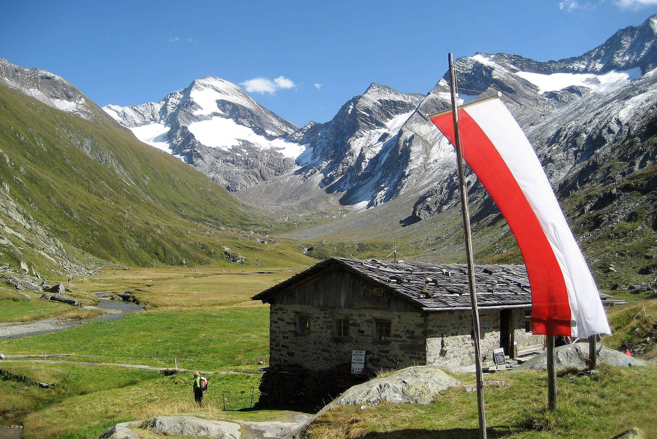 Stone cabin and Austrian flag in green alpine valley with snowy mountain peaks under a clear blue sky. - Residence Aichner