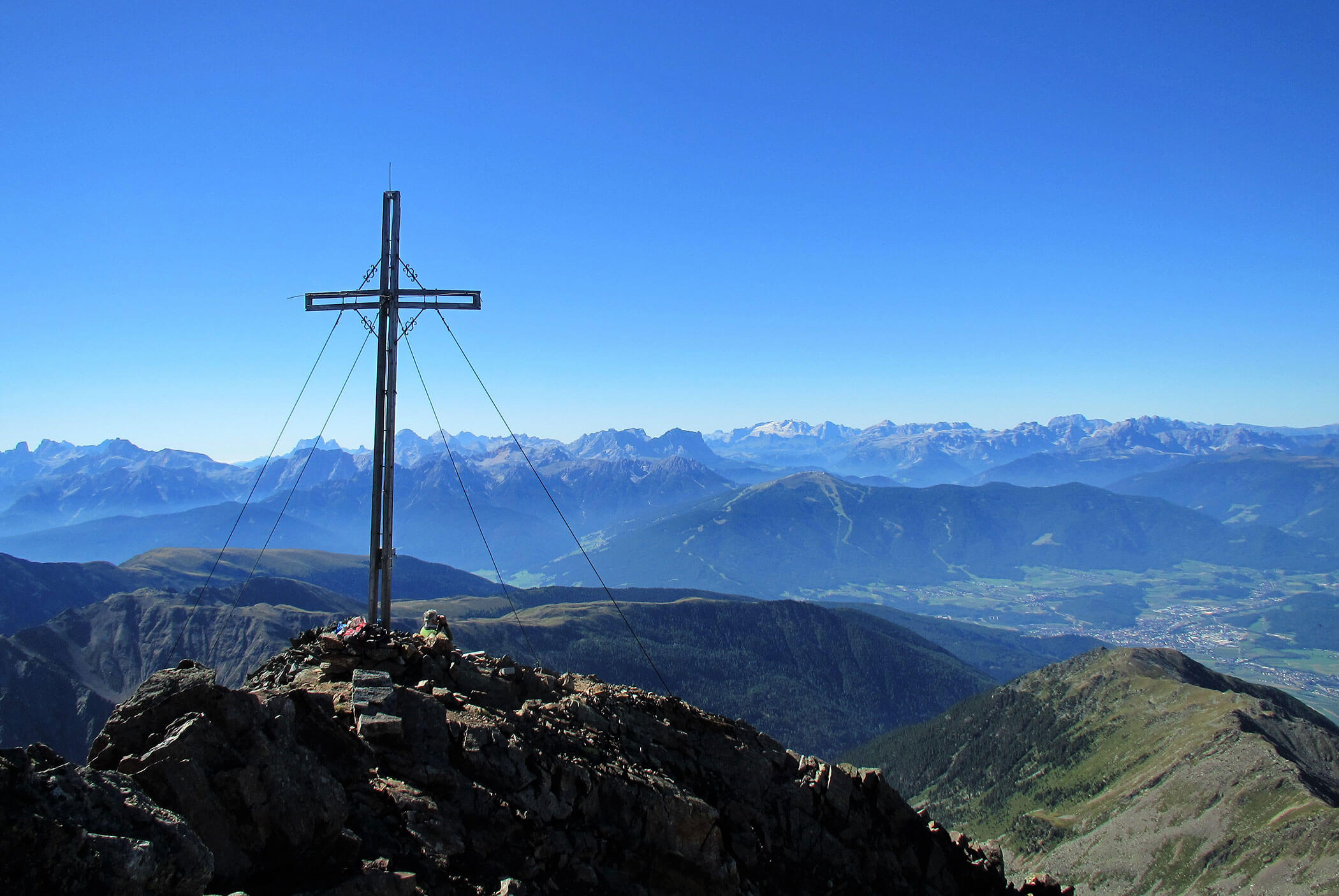 A large cross stands on a rocky mountain peak with distant mountains and valleys under a clear blue sky. - Residence Aichner