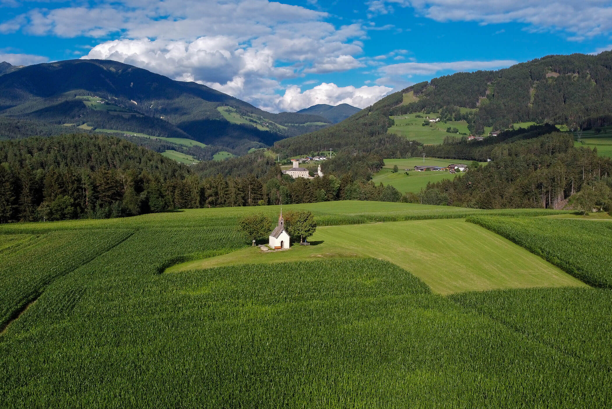 Small white chapel sits alone in a green field, surrounded by hills and mountains under a blue sky. - Residence Aichner