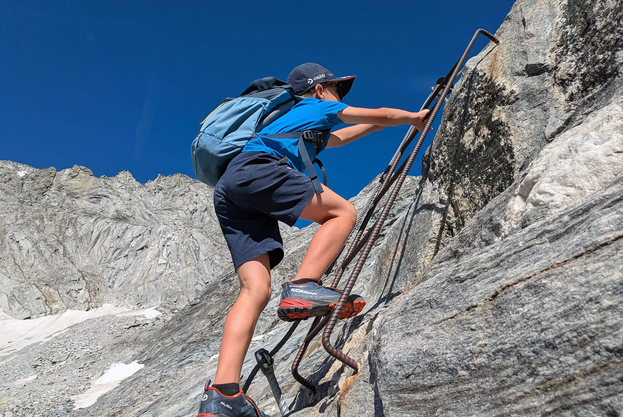A boy wearing a backpack climbs a metal ladder fixed to a steep rocky mountainside under a clear blue sky. - Residence Aichner