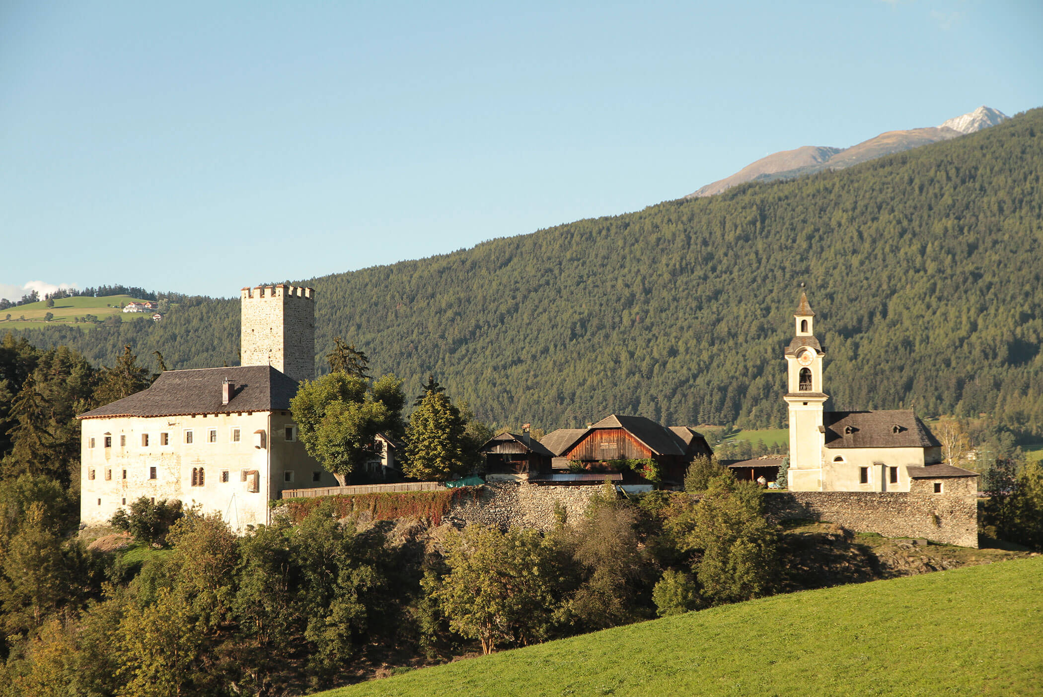 Stone castle and church on a hill, surrounded by trees and mountains under a clear sky. - Residence Aichner