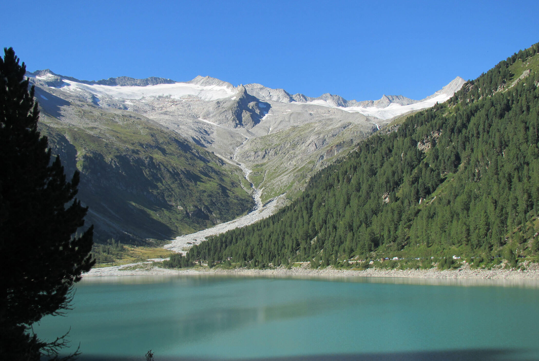 A turquoise lake with forested hillsides and snowy mountains under a clear blue sky. - Residence Aichner