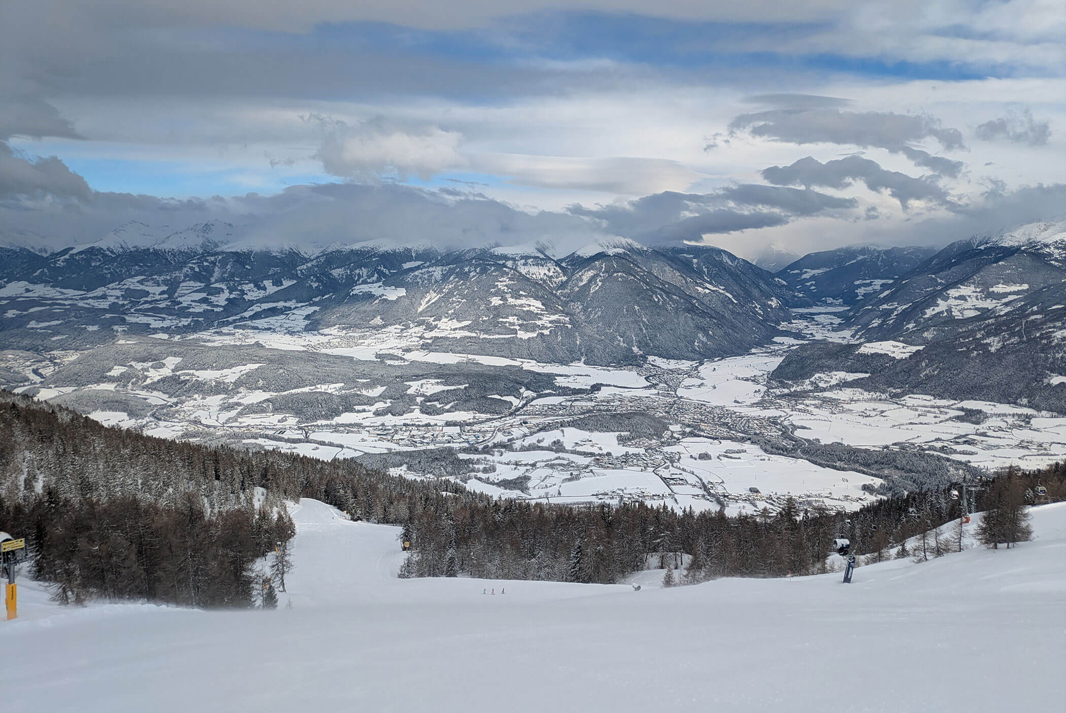 Snowy mountain landscape with scattered trees, cloudy sky, and distant valleys covered in white. - Residence Aichner