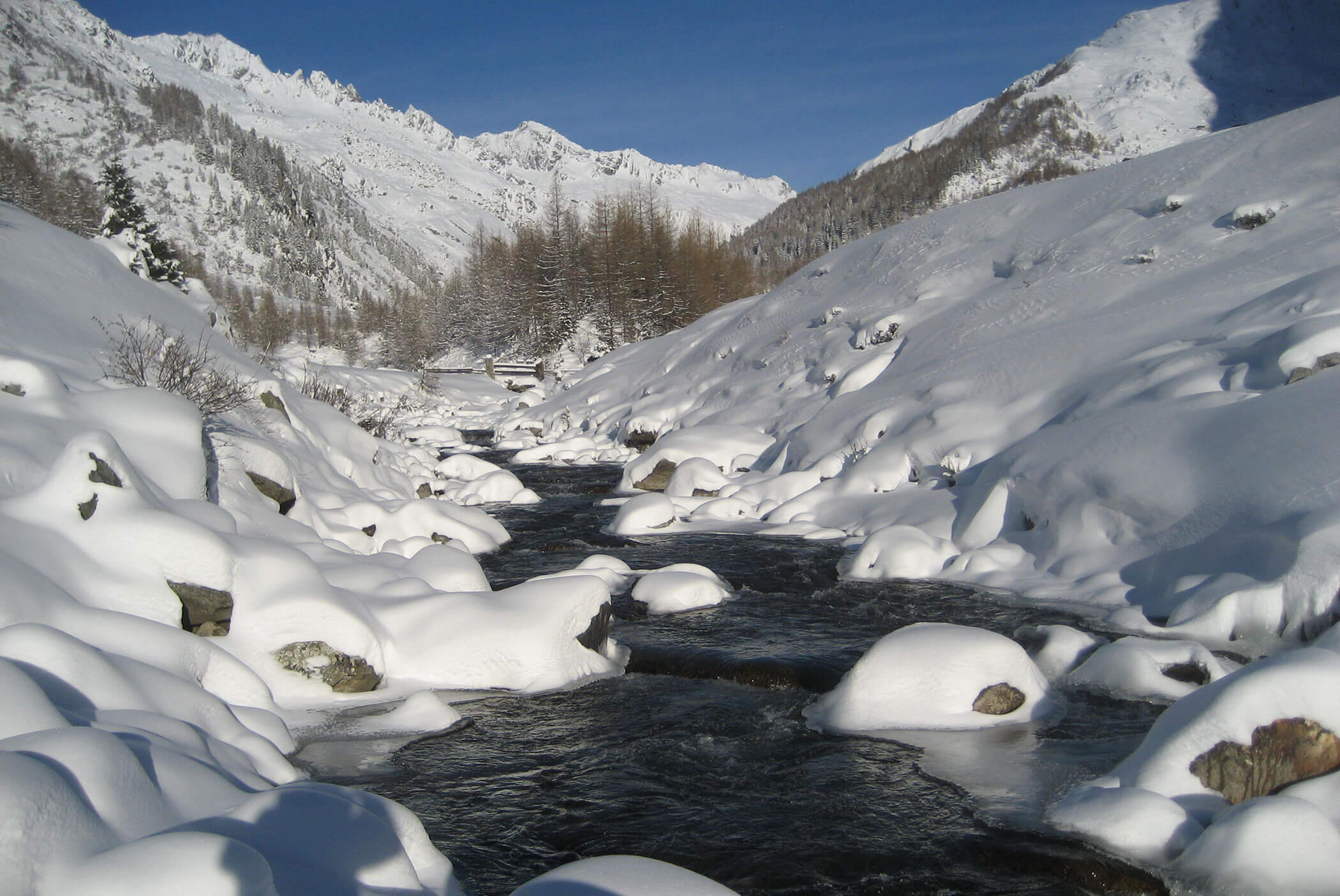 A snowy mountain landscape with a stream flowing between snow-covered rocks and trees under a blue sky. - Residence Aichner