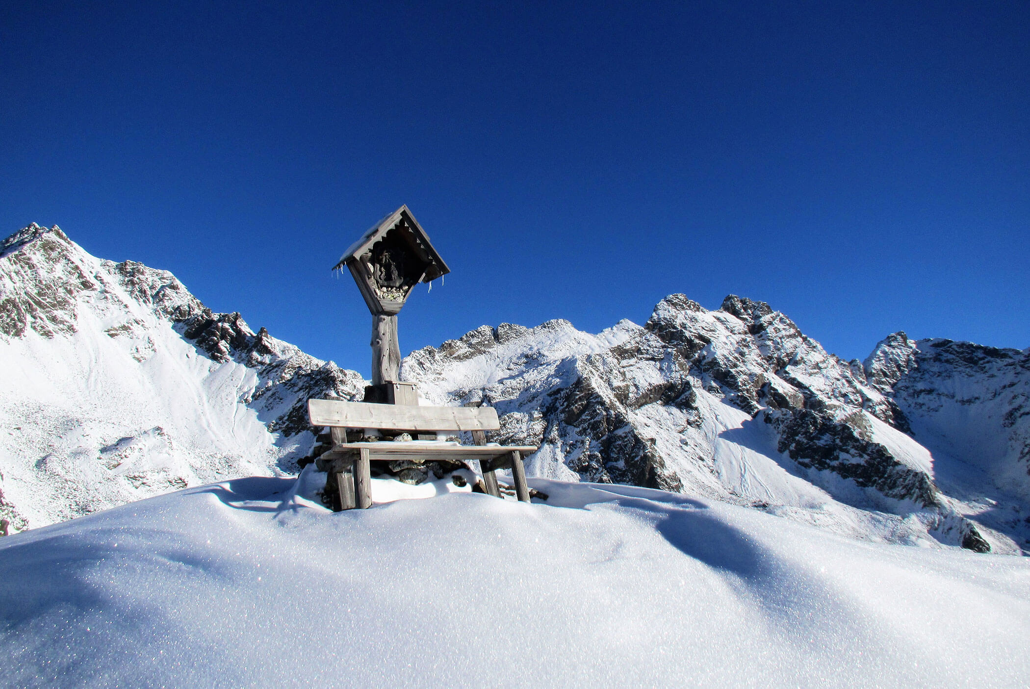 A wooden cross and bench sit in deep snow with snowy mountains and a clear blue sky in the background. - Residence Aichner