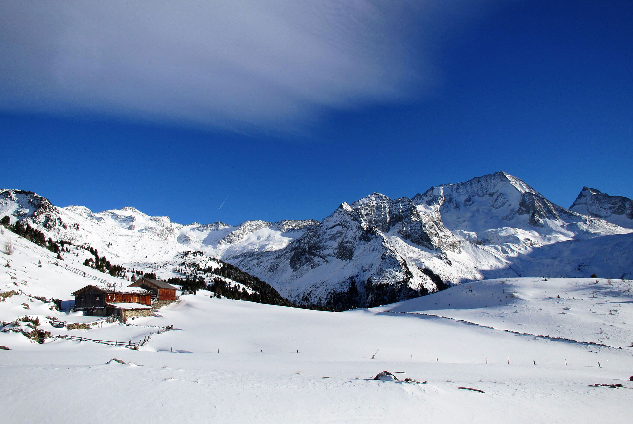 Verschneite Berge unter einem strahlend blauen Himmel, mit einer Holzhütte auf der linken Seite des Bildes. - Residence Aichner