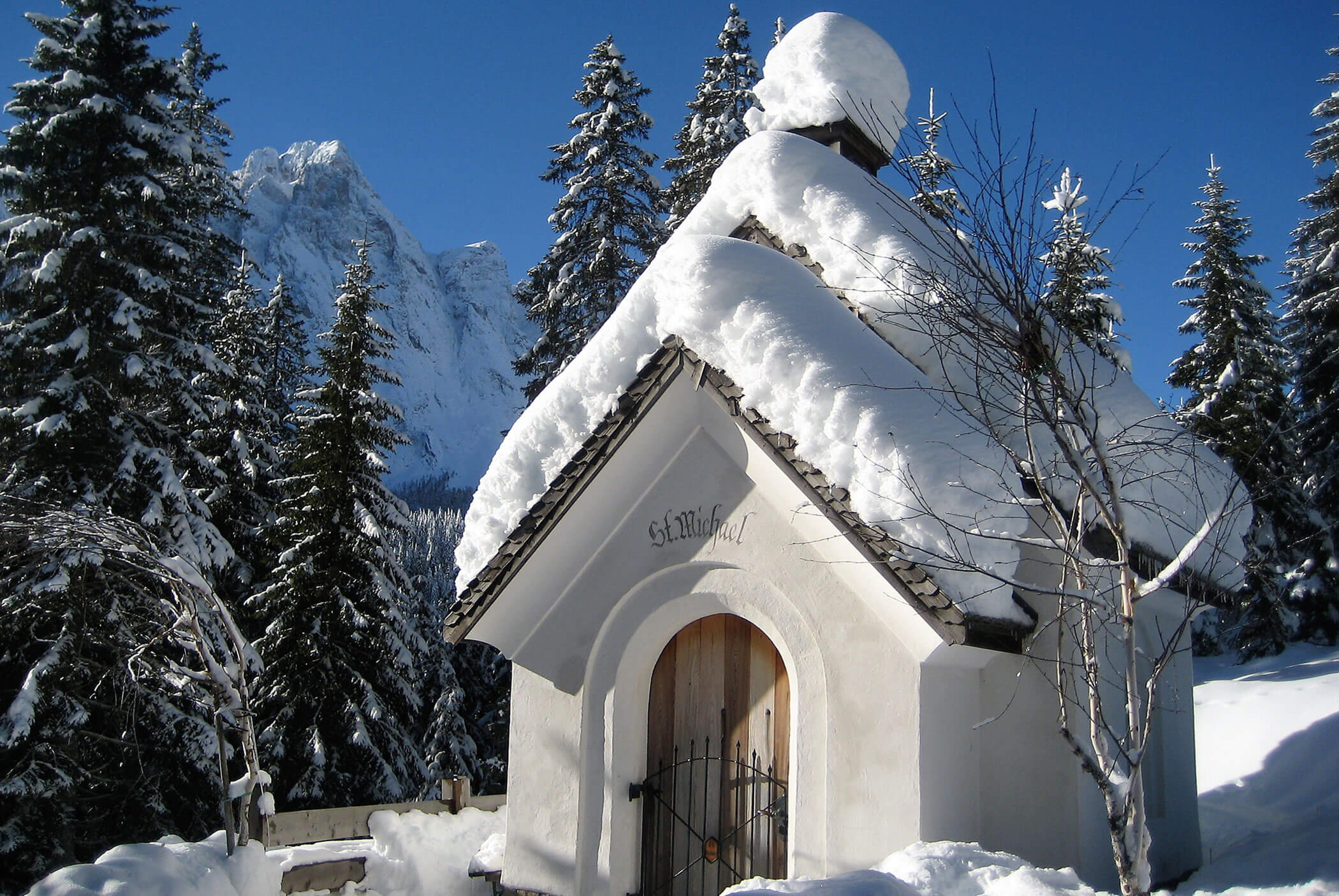 A small snow-covered chapel stands amid snowy trees with mountains in the background under a clear sky. - Residence Aichner