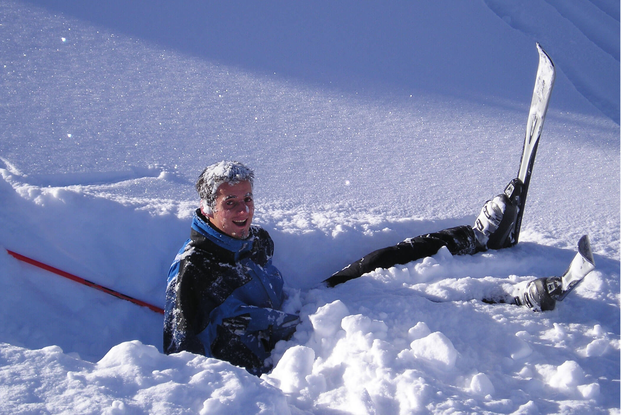 A skier sitting in deep snow, smiling, with skis sticking out at odd angles. - Residence Aichner