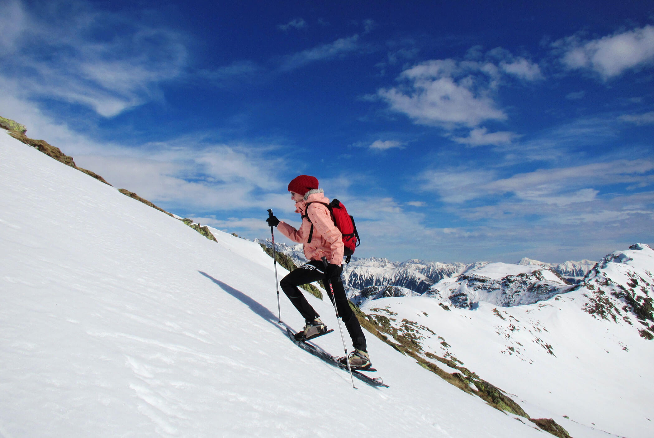 Eine Person in Winterkleidung erklimmt mit Skistöcken einen verschneiten Hang; im Hintergrund sind Berge und ein blauer Himmel zu sehen. - Residence Aichner