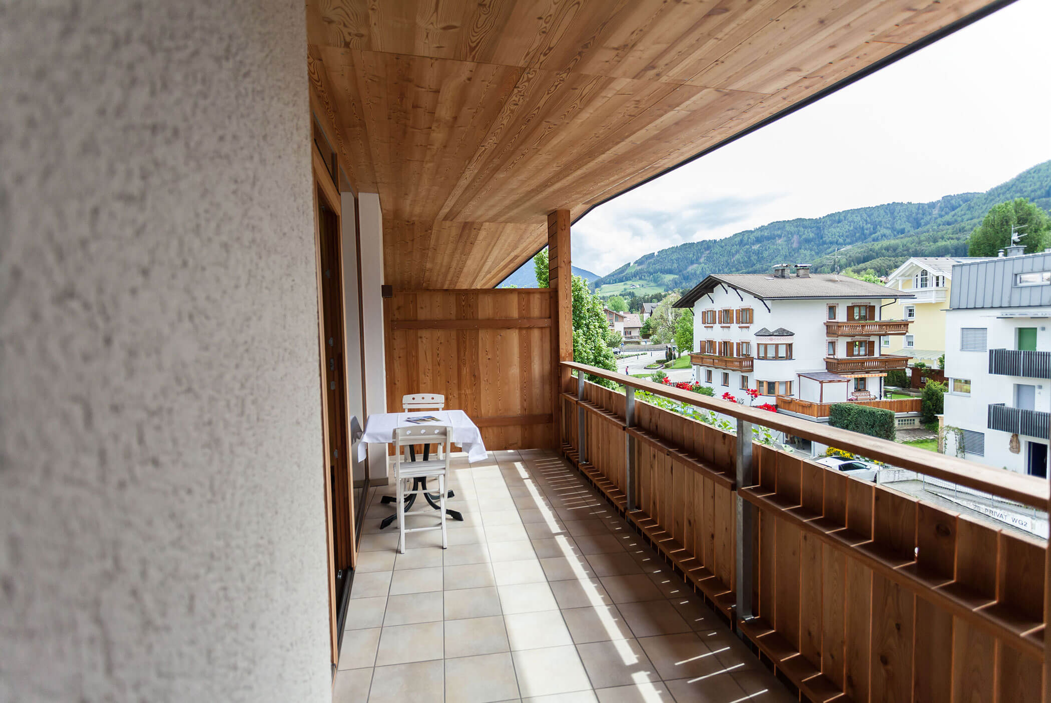 Balcone in legno con tavolo e sedie, affacciato sulle case e sulle colline verdeggianti sotto un cielo parzialmente nuvoloso. - Residence Aichner