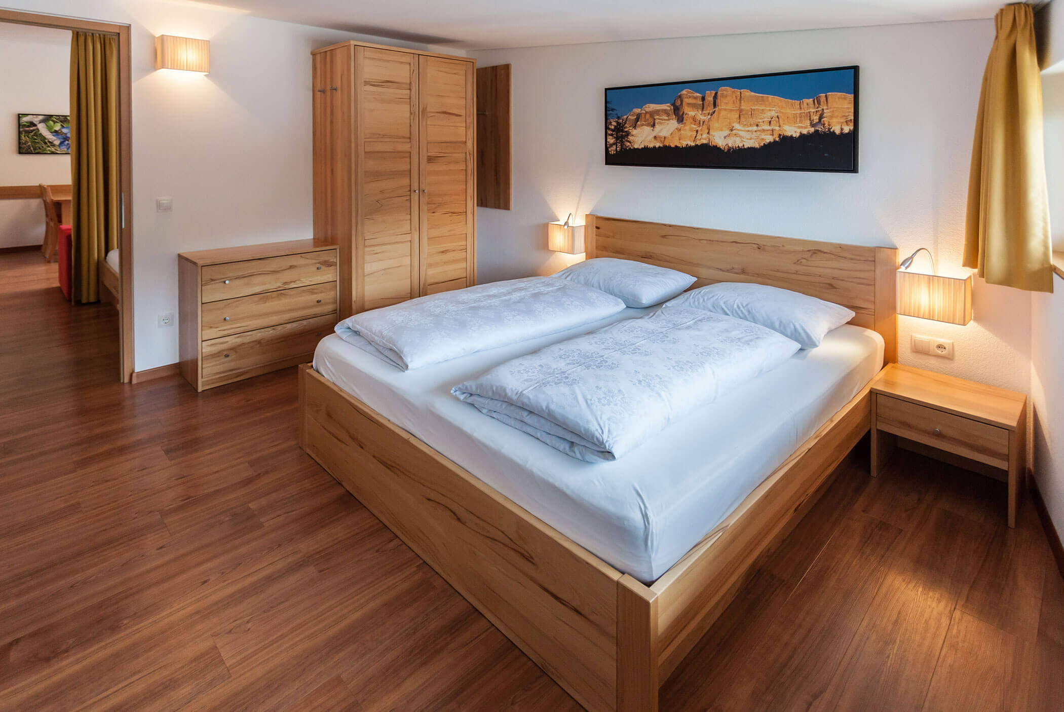 Camera da letto luminosa con mobili in legno, un quadro sopra il letto e la luce del sole che filtra attraverso le tende gialle. - Residence Aichner