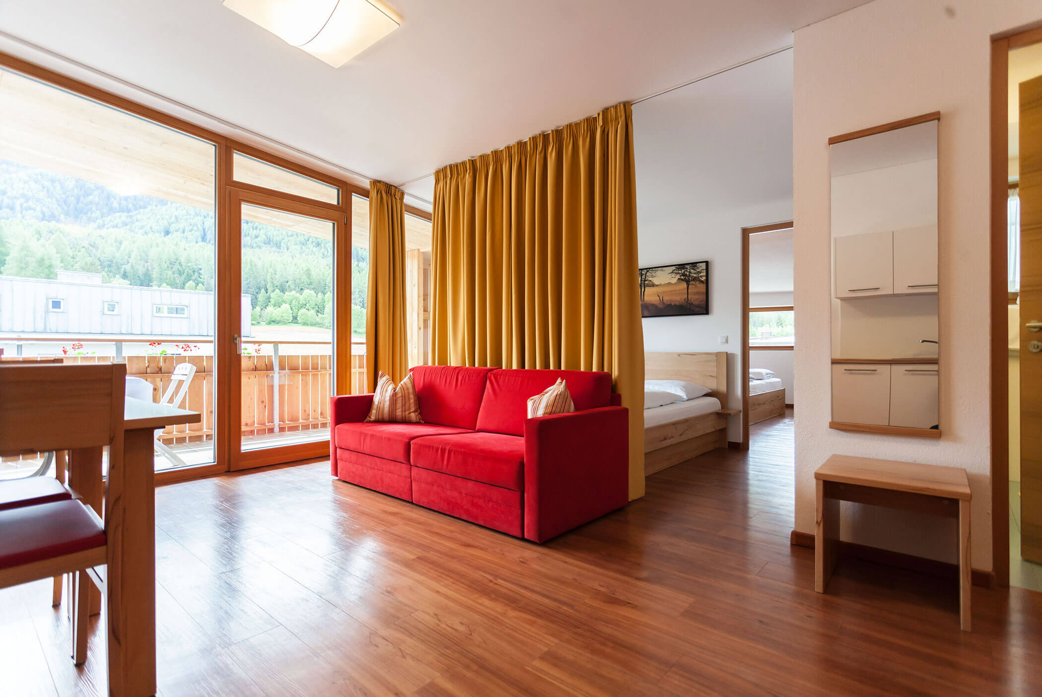 Luminosa zona giorno con divano rosso, pavimento in legno, tenda gialla, balcone e vista su una camera da letto. - Residence Aichner