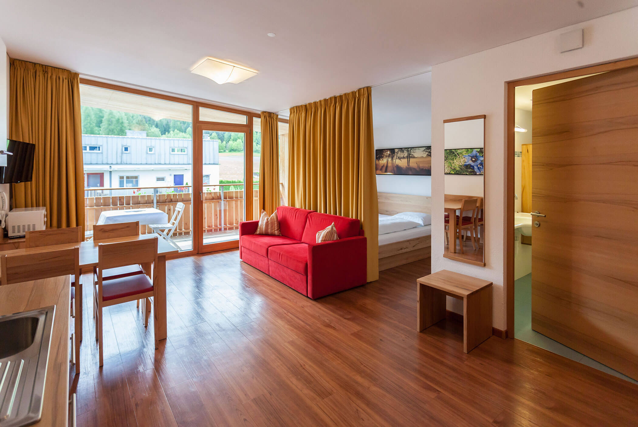 Luminoso appartamento con pavimenti in legno, divano rosso, tavolo da pranzo, balcone e una camera da letto parzialmente visibile attraverso una porta. - Residence Aichner