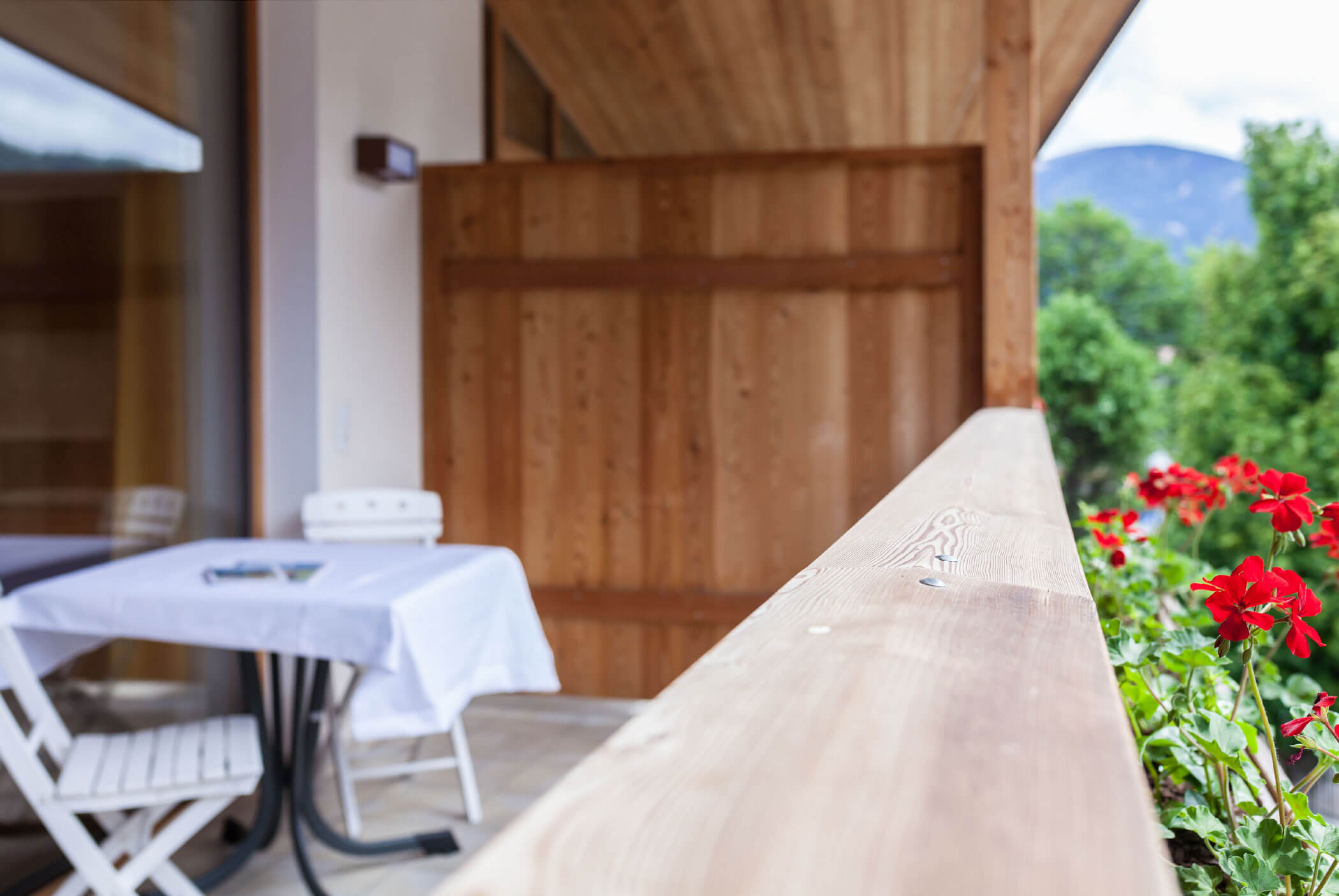 Balcone in legno con un tavolo e delle sedie bianche, fiori rossi e alberi verdi sullo sfondo. - Residence Aichner