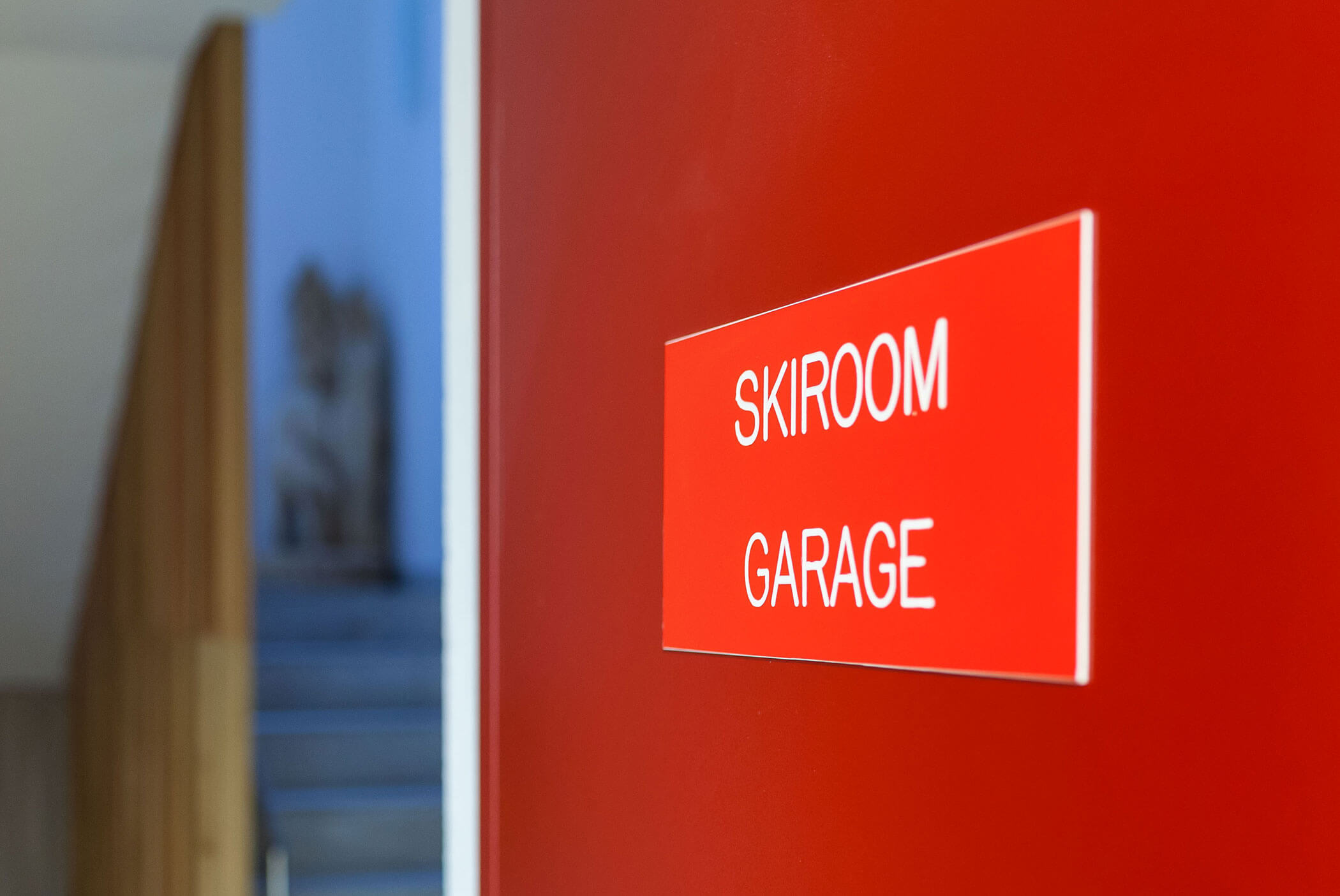 Rote Tür mit einem Schild, auf dem in weißen Buchstaben „SKIROOM GARAGE“ steht; im Hintergrund eine verschwommene Treppe. - Residence Aichner