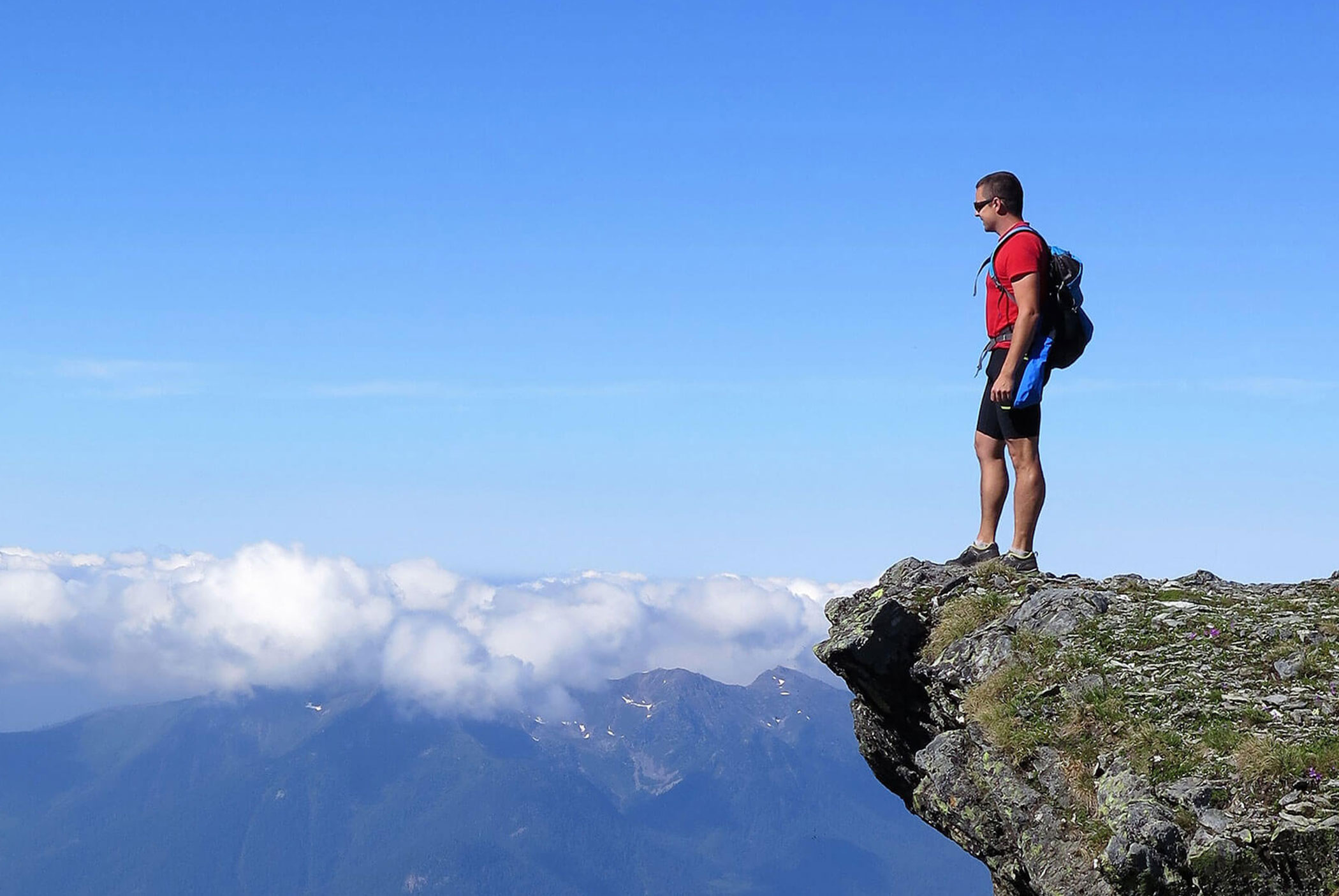 Ein Wanderer steht am Rand einer felsigen Klippe und blickt unter blauem Himmel auf Berge und Wolken. - Residence Aichner