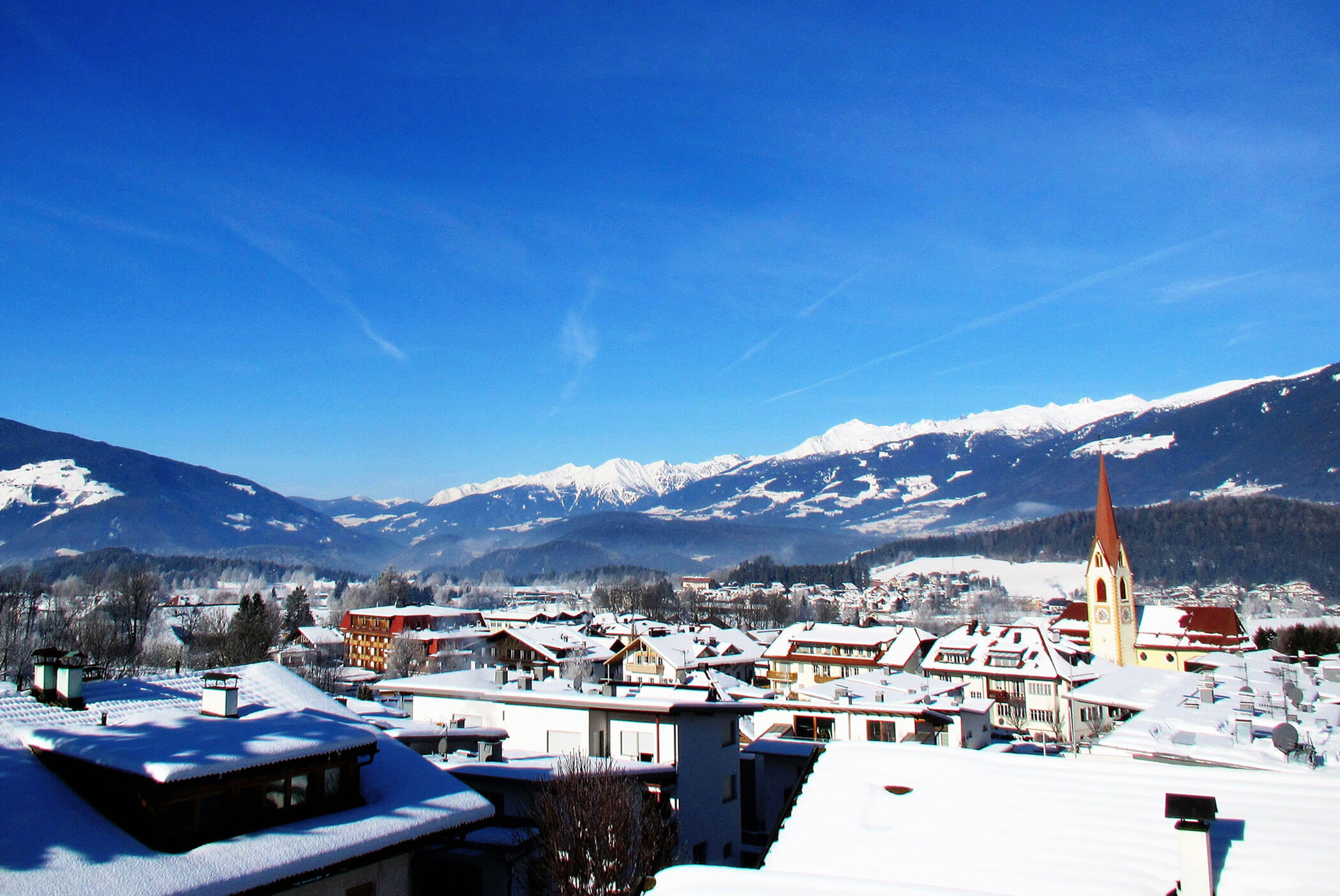 Ein schneebedecktes Dorf mit Bergen im Hintergrund unter strahlend blauem Himmel. Der Kirchturm sticht hervor. - Residence Aichner