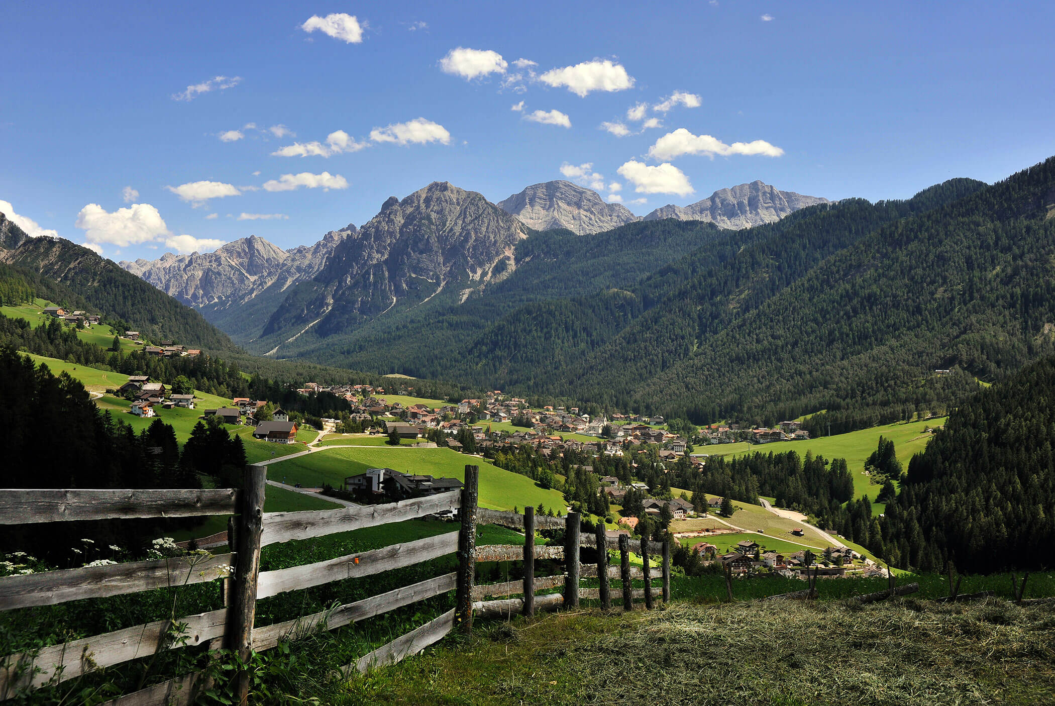Ein Holzzaun bietet den Blick auf ein Dorf, das in einem grünen Tal liegt und von hohen, bewaldeten Bergen umgeben ist. - Residence Aichner
