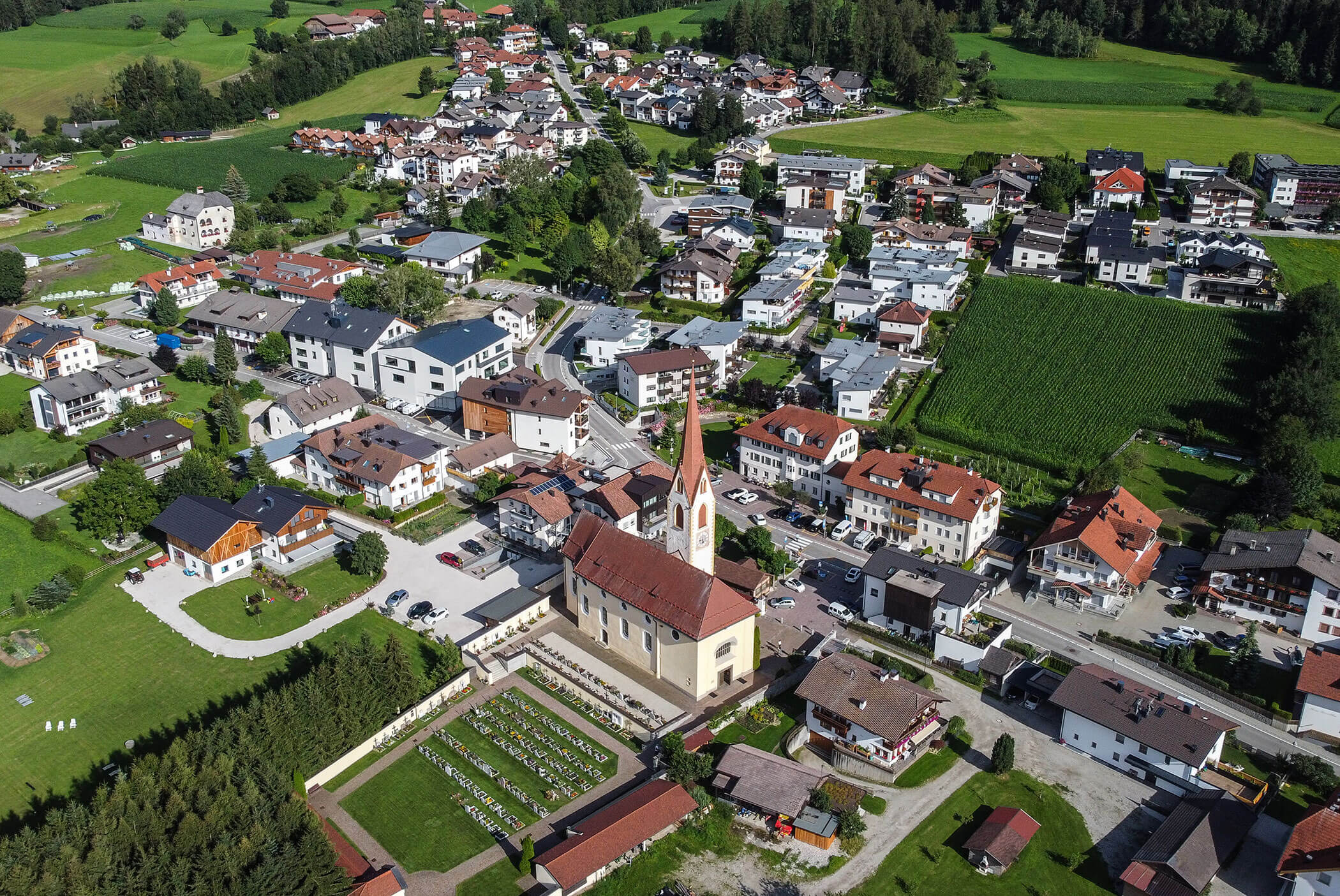 Luftaufnahme eines Dorfes mit einer Kirche, Häusern, grünen Feldern und umliegenden Bäumen an einem sonnigen Tag. - Residence Aichner