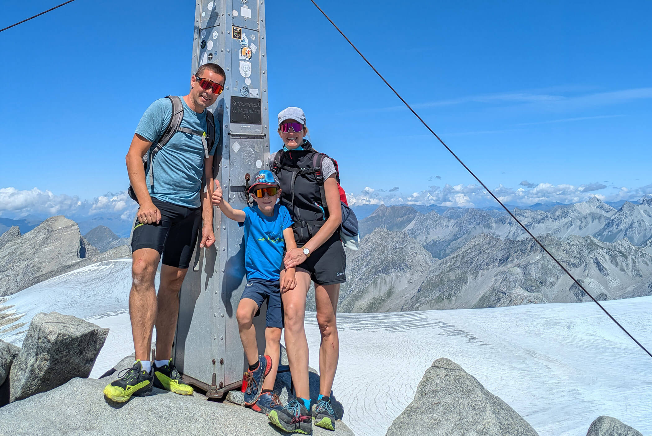 Drei Personen in Wanderkleidung posieren neben einem Gipfelstein auf einem schneebedeckten Berg, im Hintergrund sind felsige Gipfel zu sehen. - Residence Aichner