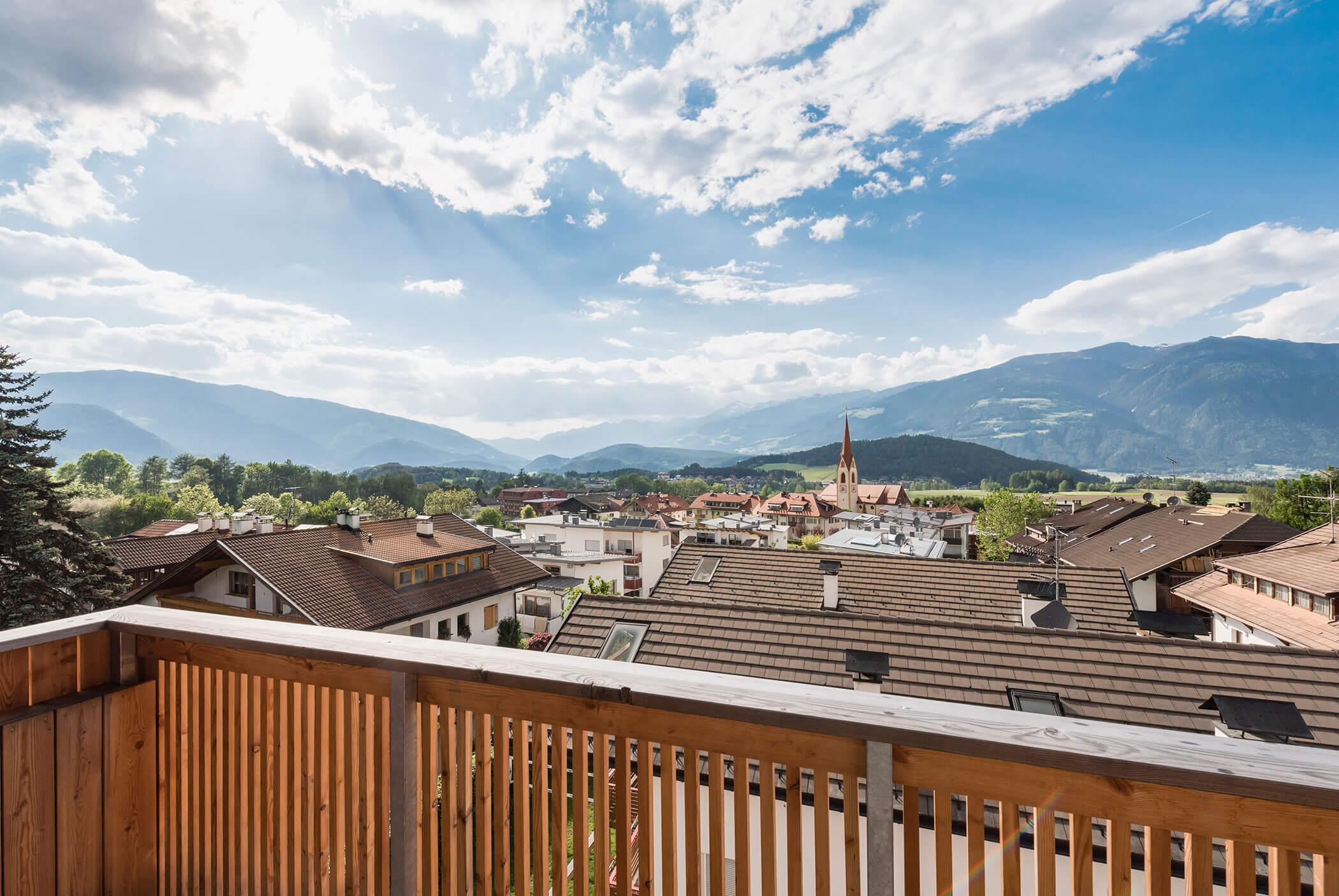 Ein Holzbalkon mit Blick auf die Dächer, eine Kirche, grüne Bäume und ferne Berge unter einem teilweise bewölkten Himmel. - Residence Aichner