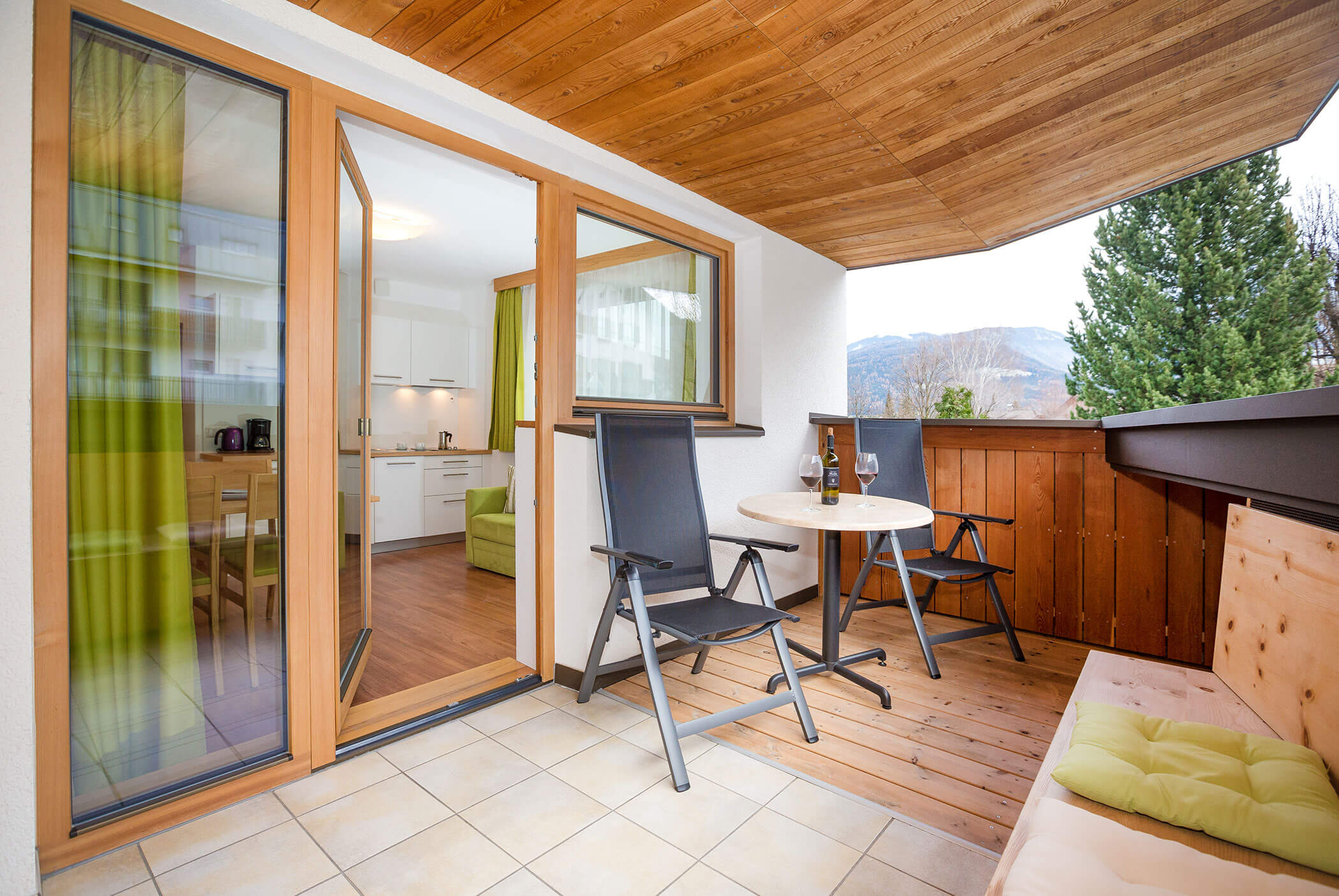 Überdachter Balkon mit zwei Stühlen, einem Tisch und einer Bank, mit Blick auf Bäume und Berge, direkt neben der Küche. - Residence Aichner