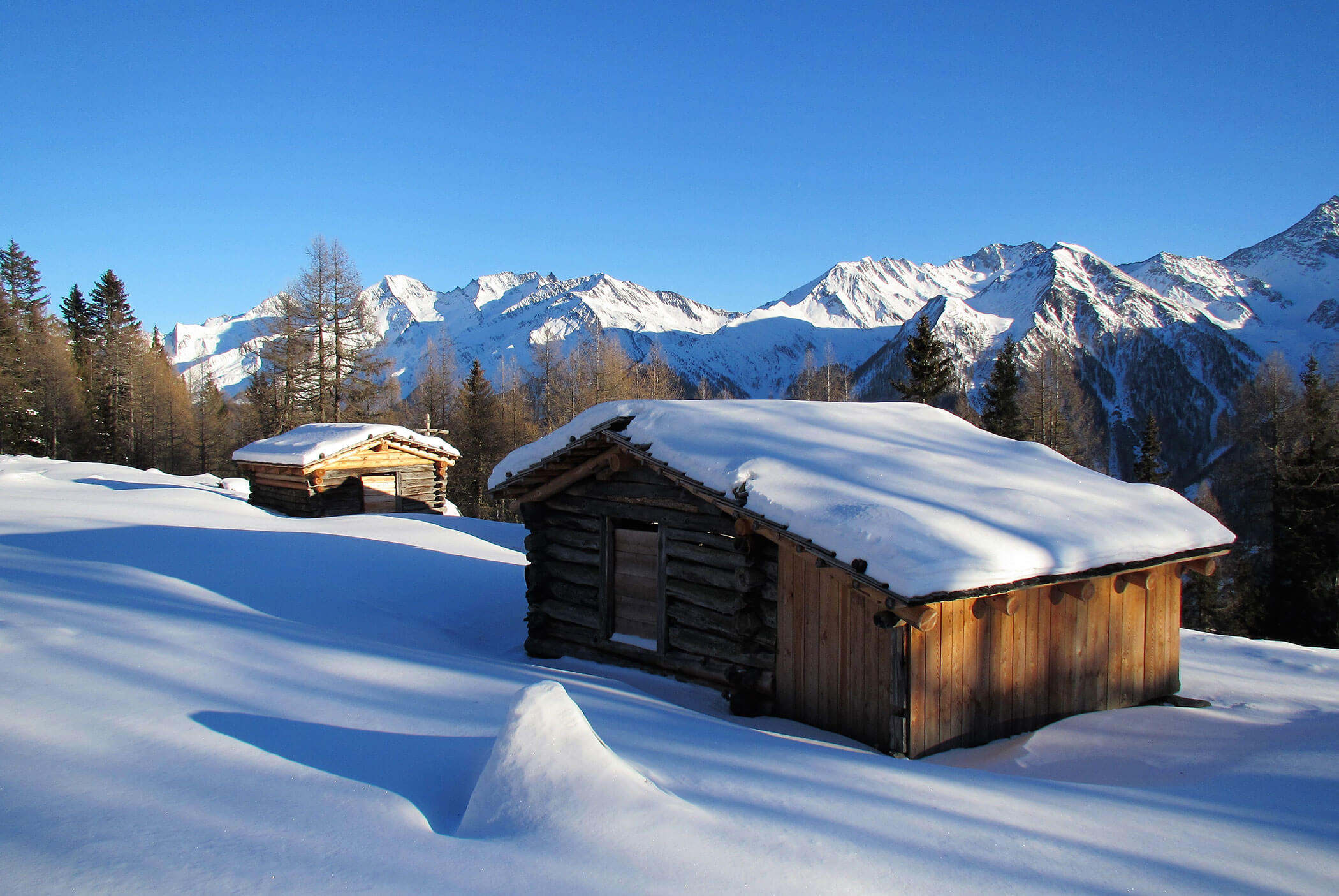 Zwei Holzhütten stehen im tiefen Schnee, im Hintergrund sind Berggipfel und ein strahlend blauer Himmel zu sehen. - Residence Aichner