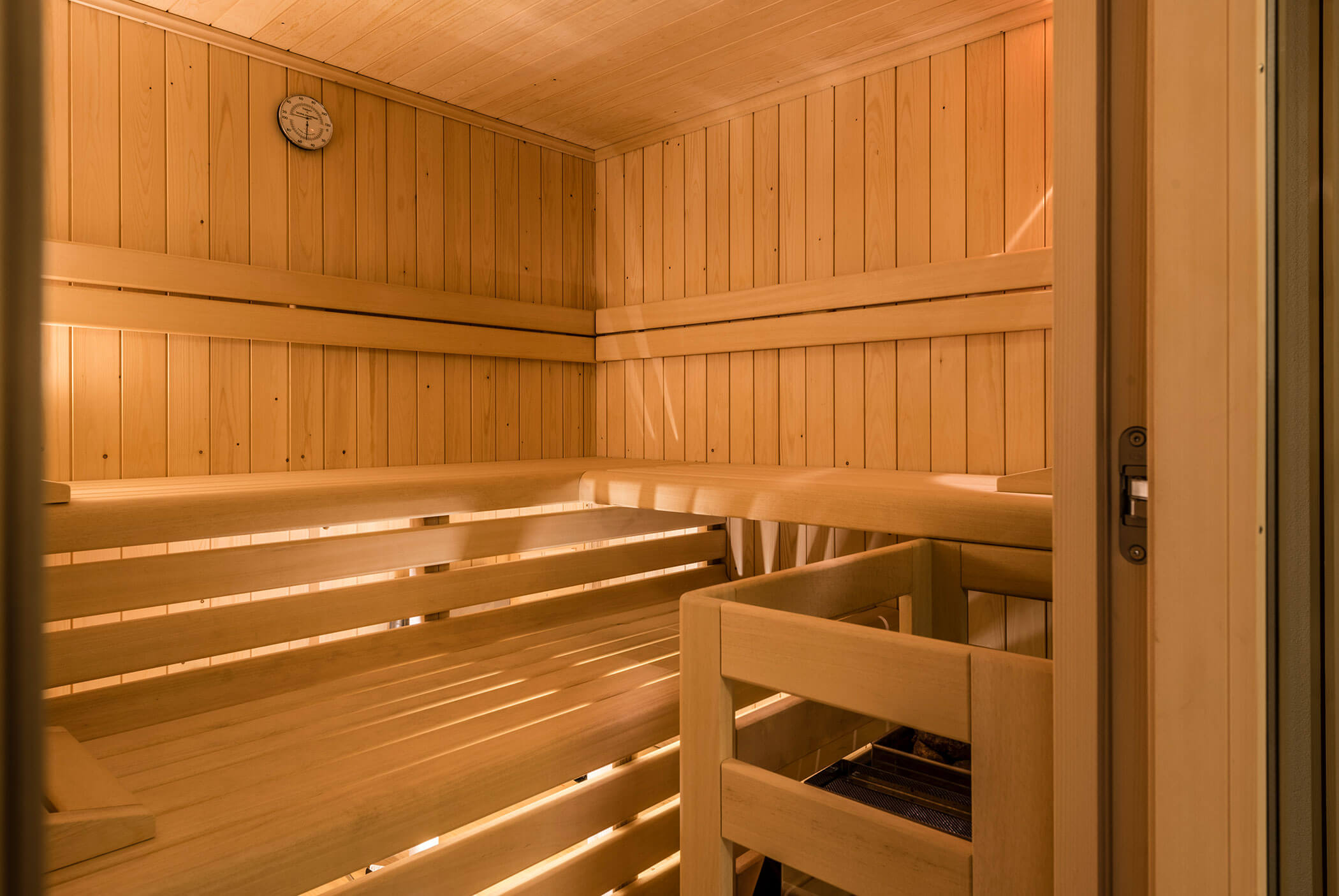 Eine Holzsauna mit Sitzbänken und einer Wanduhr, die in warmes, sanftes gelbes Licht getaucht ist. - Residence Aichner