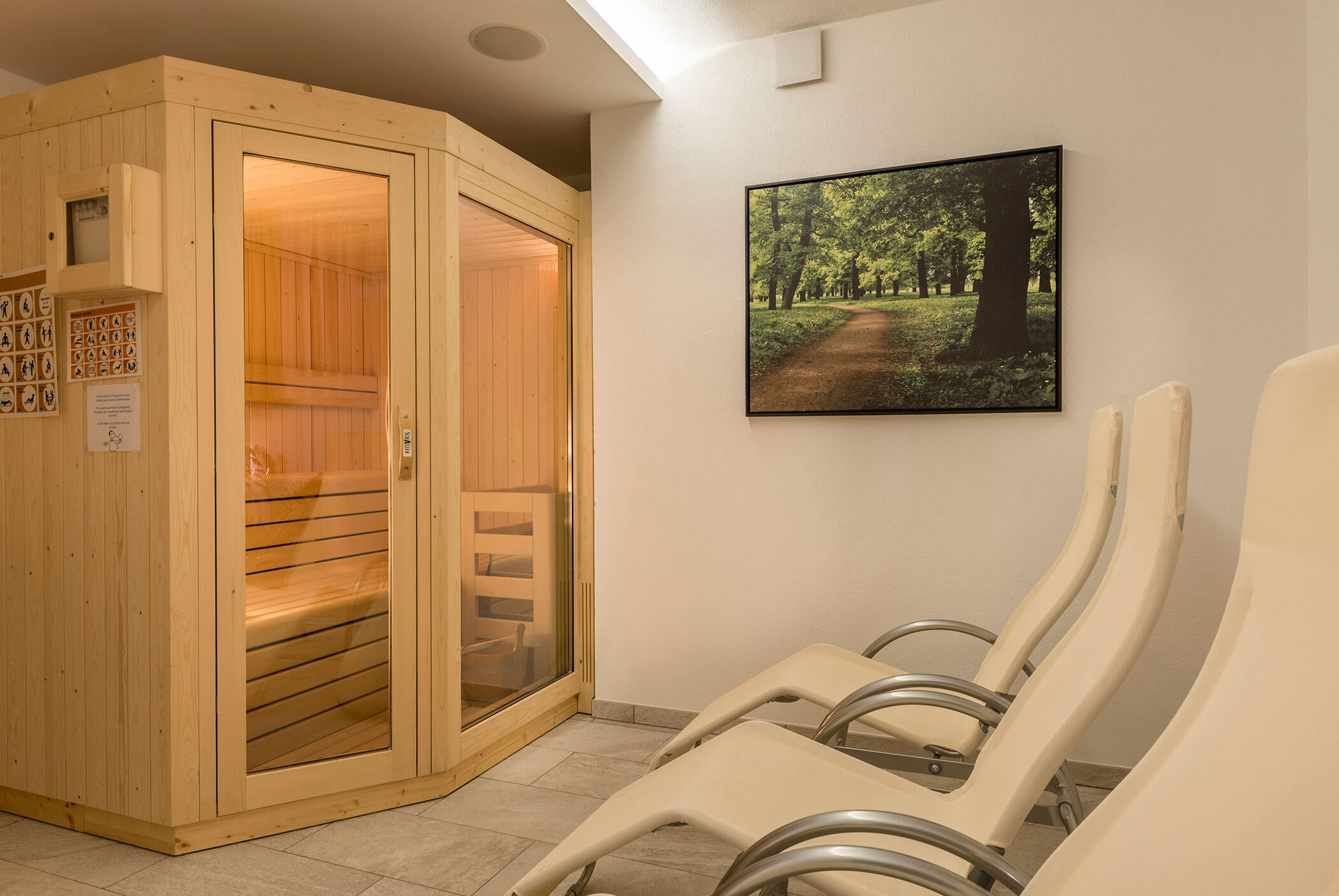 Wood sauna and three cream lounge chairs in a clean room, with a large framed photo of a tree-lined path. - Residence Aichner