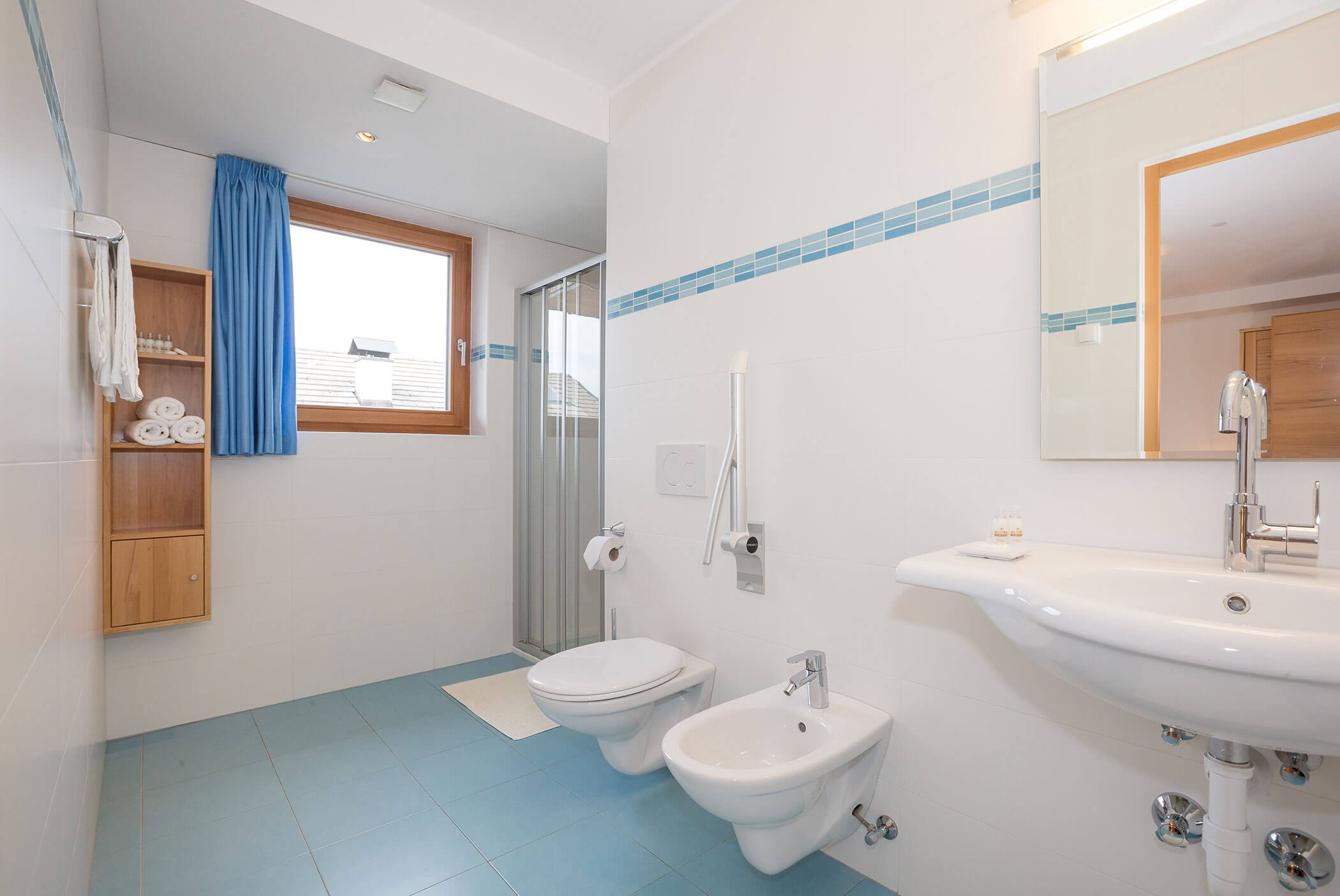 Bagno luminoso con pavimento in piastrelle blu, lavabo sospeso, WC, bidet, doccia, finestra e mensole. - Residence Aichner