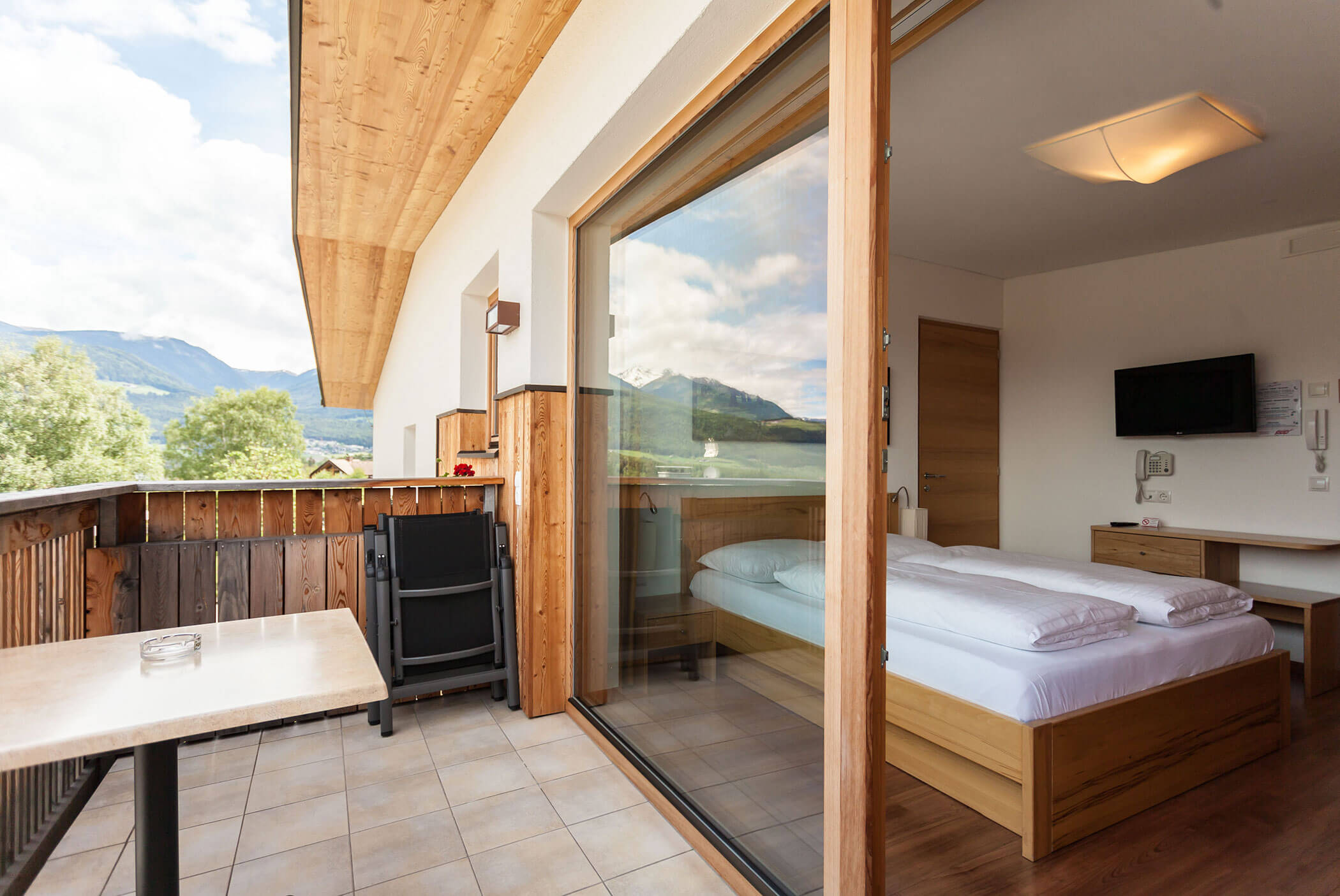Il balcone dell'hotel, arredato con tavolo e sedie, si affaccia sulle montagne; la porta a vetri conduce a una camera da letto ordinata, dotata di TV e letto. - Residence Aichner