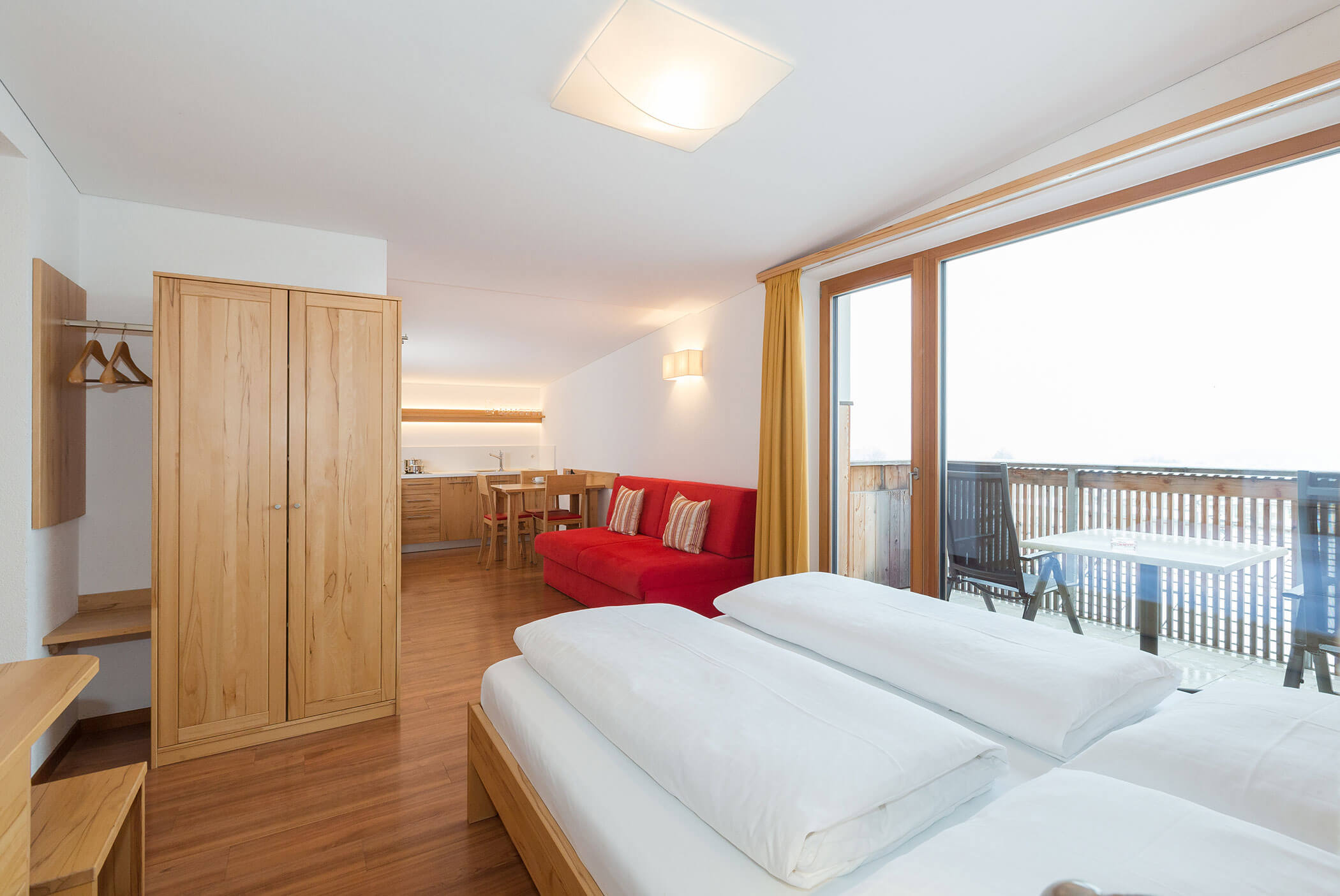 Camera d'albergo luminosa con letto matrimoniale, divano rosso, armadio, tavolo e balcone con posti a sedere all'aperto. - Residence Aichner