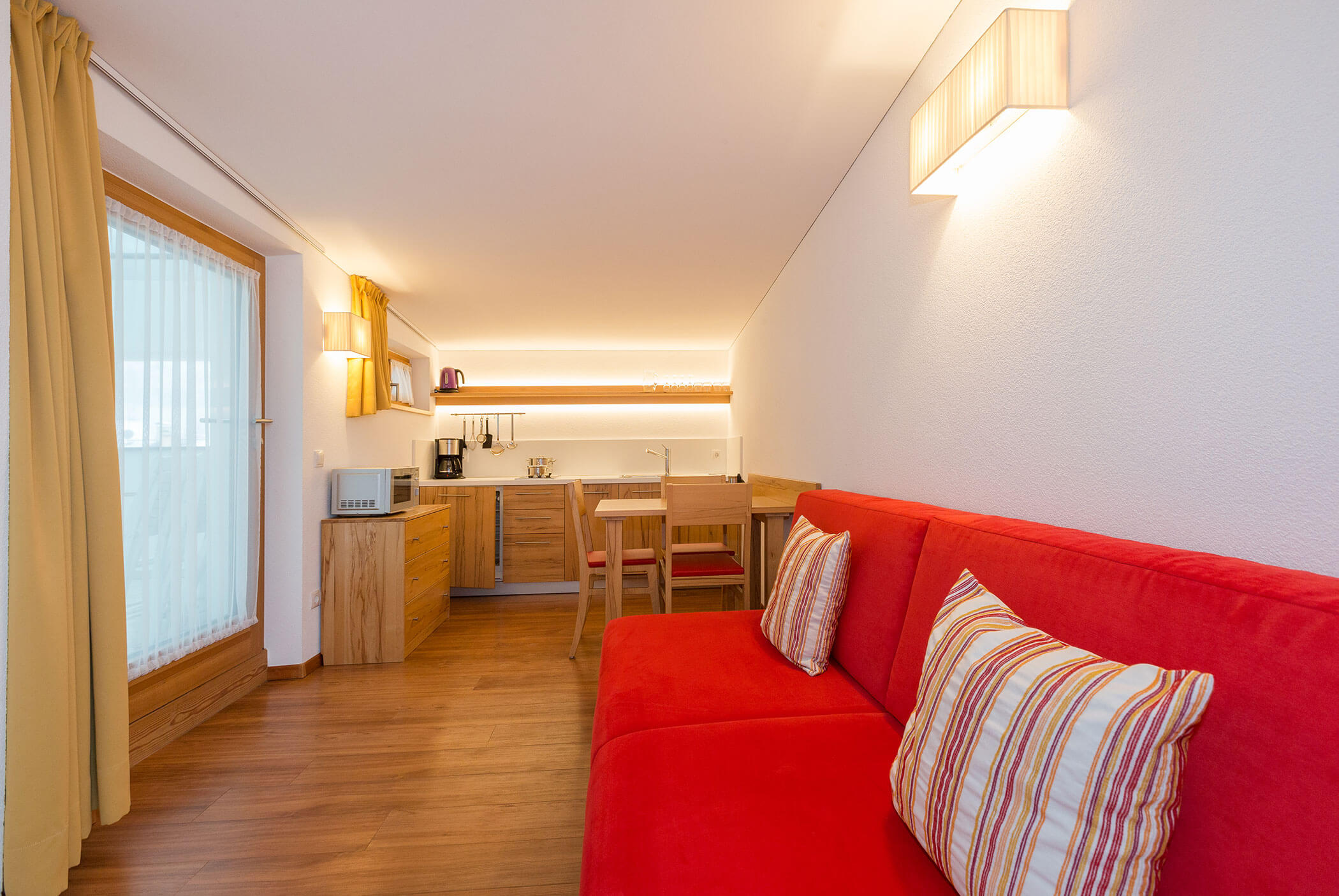 Moderno monolocale con divano rosso, cuscini a righe, angolo cottura, armadi in legno e illuminazione brillante. - Residence Aichner