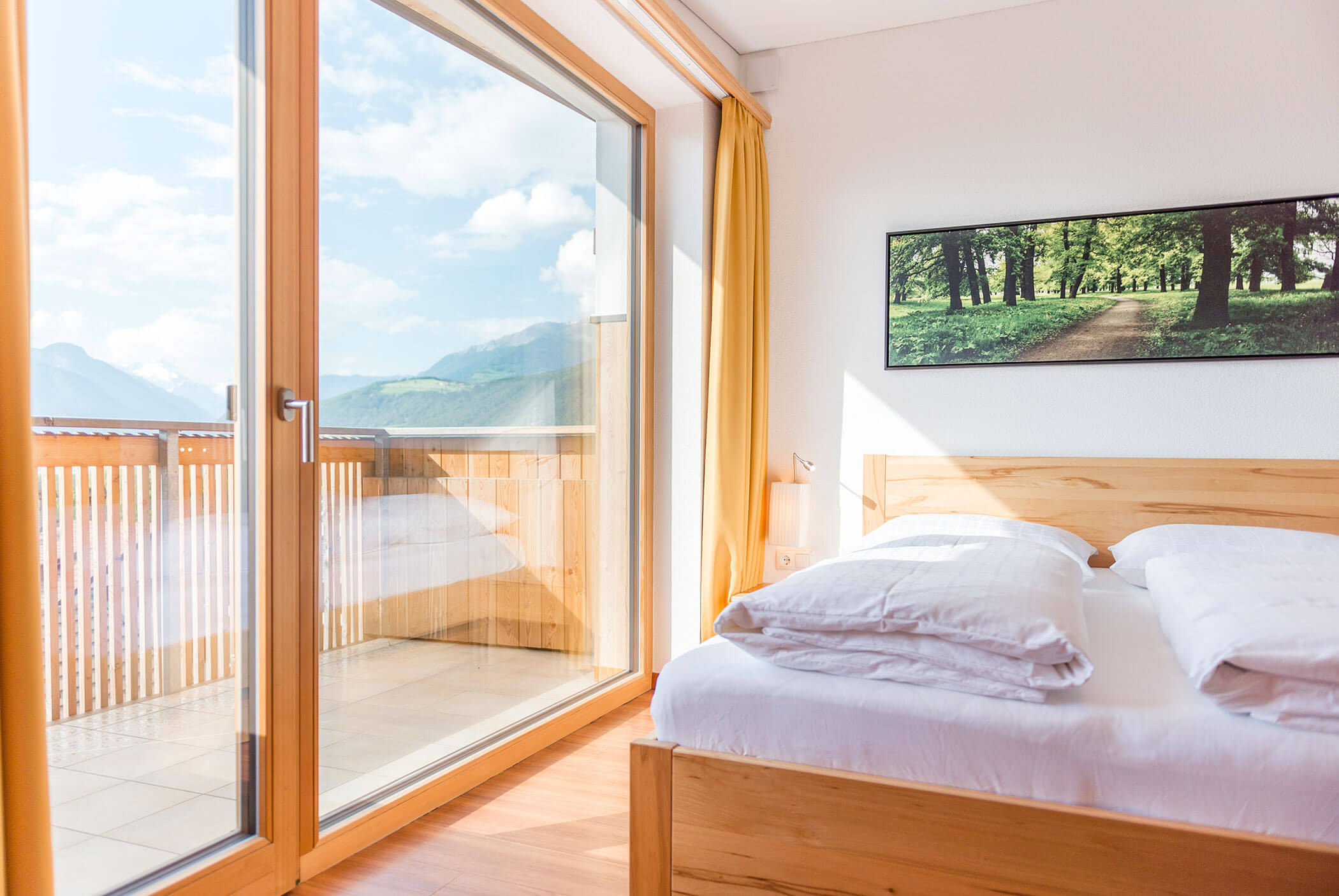 Camera da letto luminosa con letto in legno, ampia finestra, balcone con vista sulle montagne e una foto incorniciata del parco appesa alla parete. - Residence Aichner