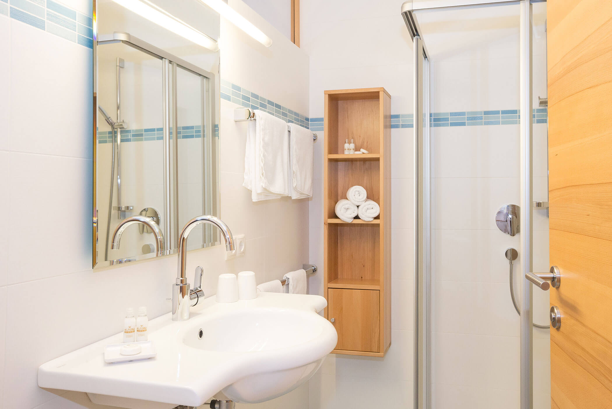 Bagno moderno con lavabo bianco, specchio, box doccia in vetro e mensole in legno con asciugamani arrotolati con cura. - Residence Aichner