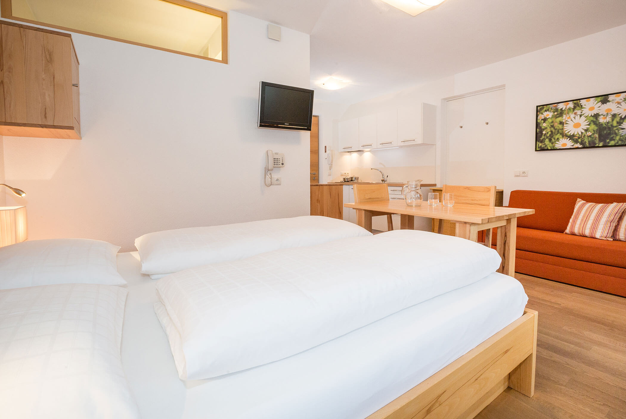 Luminoso monolocale con letto, angolo cottura, tavolo da pranzo, divano arancione e TV a parete. - Residence Aichner
