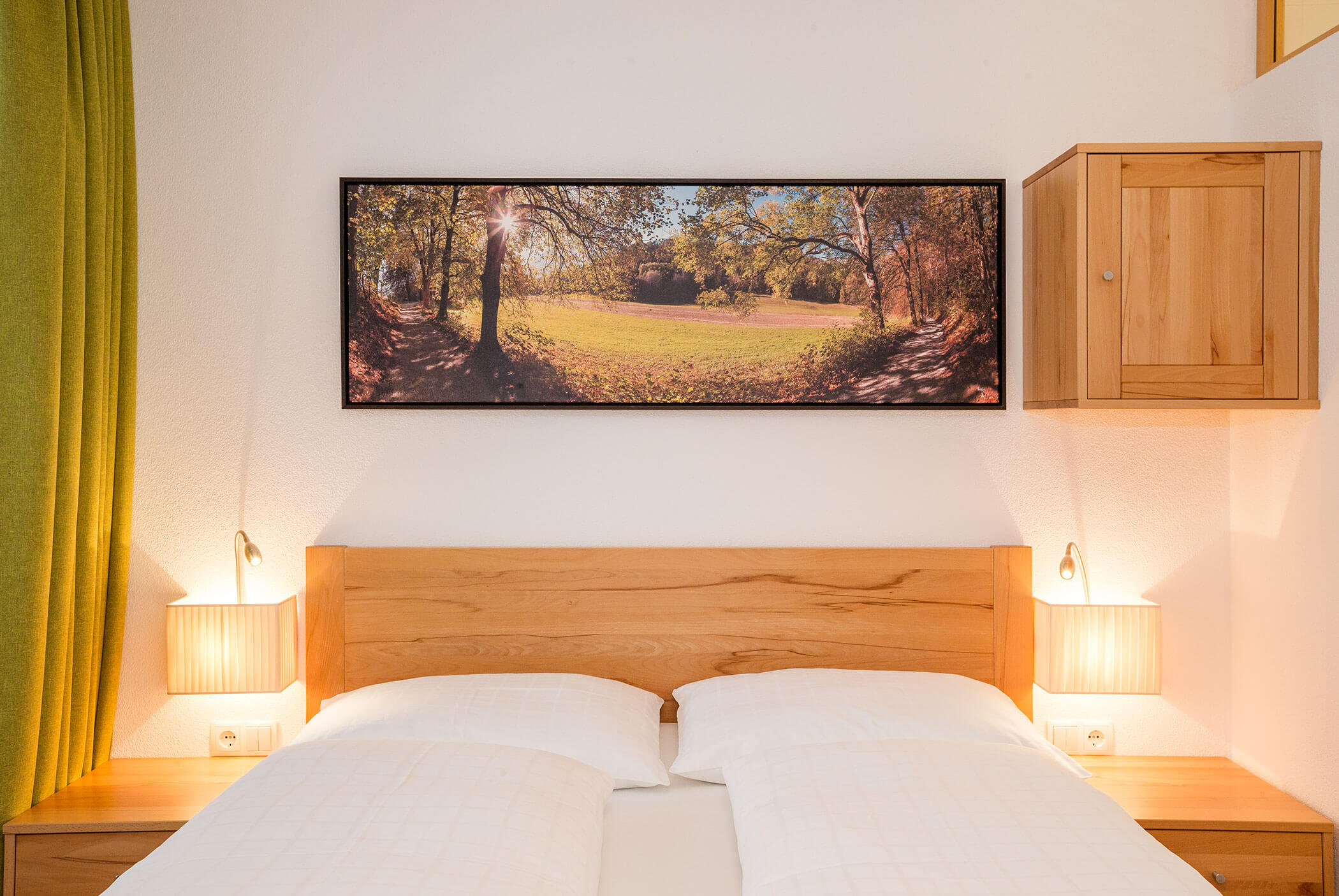 Un letto ben rifatto con lenzuola bianche, testiera in legno, due lampade da comodino e una foto di un paesaggio appesa sopra. - Residence Aichner