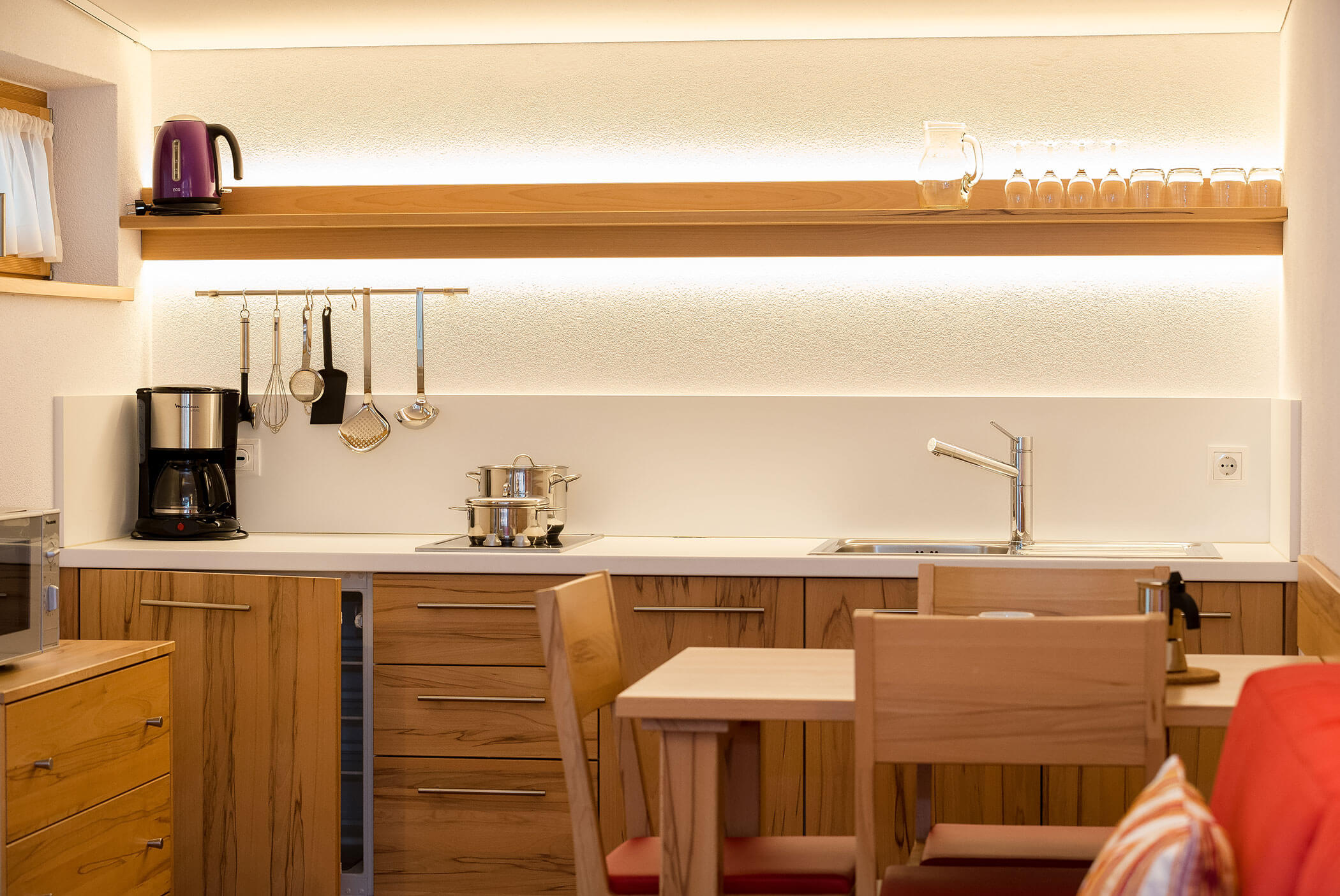 Cucina moderna con mobili in legno, macchina per il caffè, utensili, pentole, lavello e tazze su una mensola illuminata. - Residence Aichner