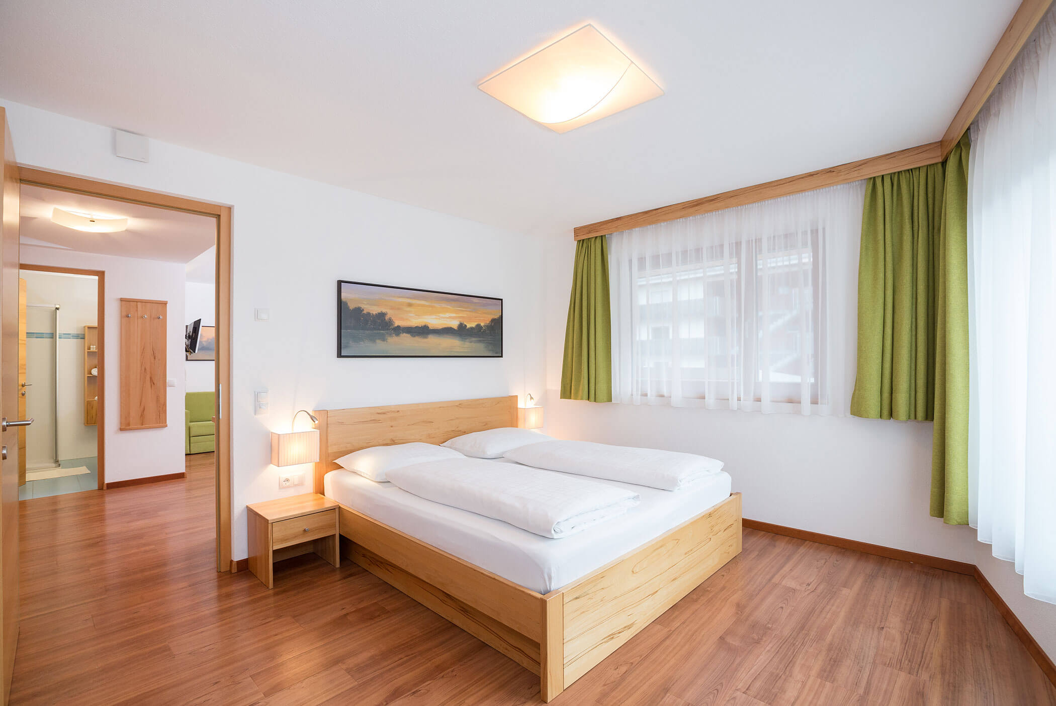 Una luminosa camera d'albergo con pavimenti in legno, letto matrimoniale, tende verdi e un quadro raffigurante un tramonto appeso alla parete. - Residence Aichner