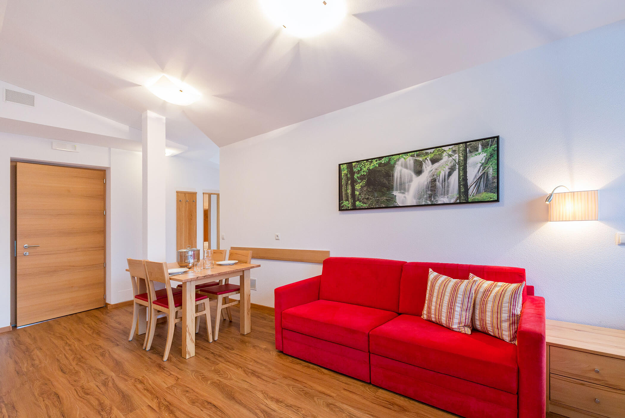 Una stanza luminosa con un divano rosso, un tavolo da pranzo, il pavimento in legno e una foto di una cascata appesa alla parete. - Residence Aichner