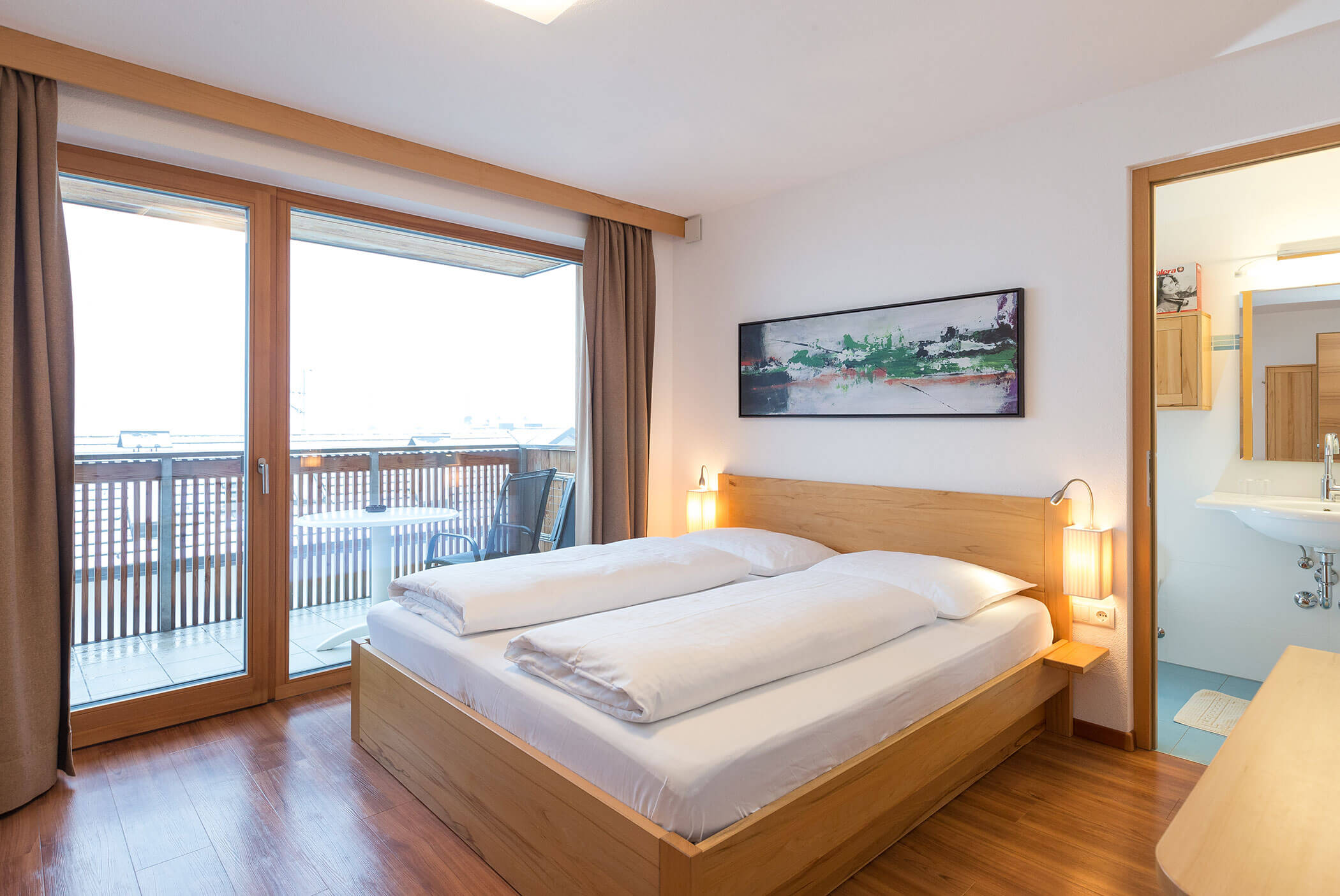 Una camera d'albergo luminosa con letto matrimoniale, accesso al balcone, pavimenti in legno e un quadro appeso sopra il letto. - Residence Aichner