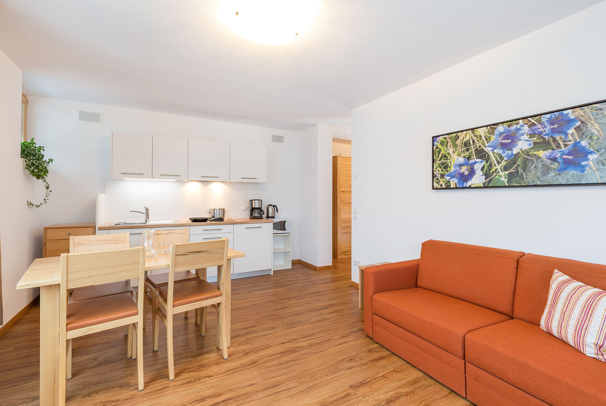 Cucina moderna e zona pranzo con pavimento in legno, divano arancione, quadri alle pareti e mobili bianchi. - Residence Aichner
