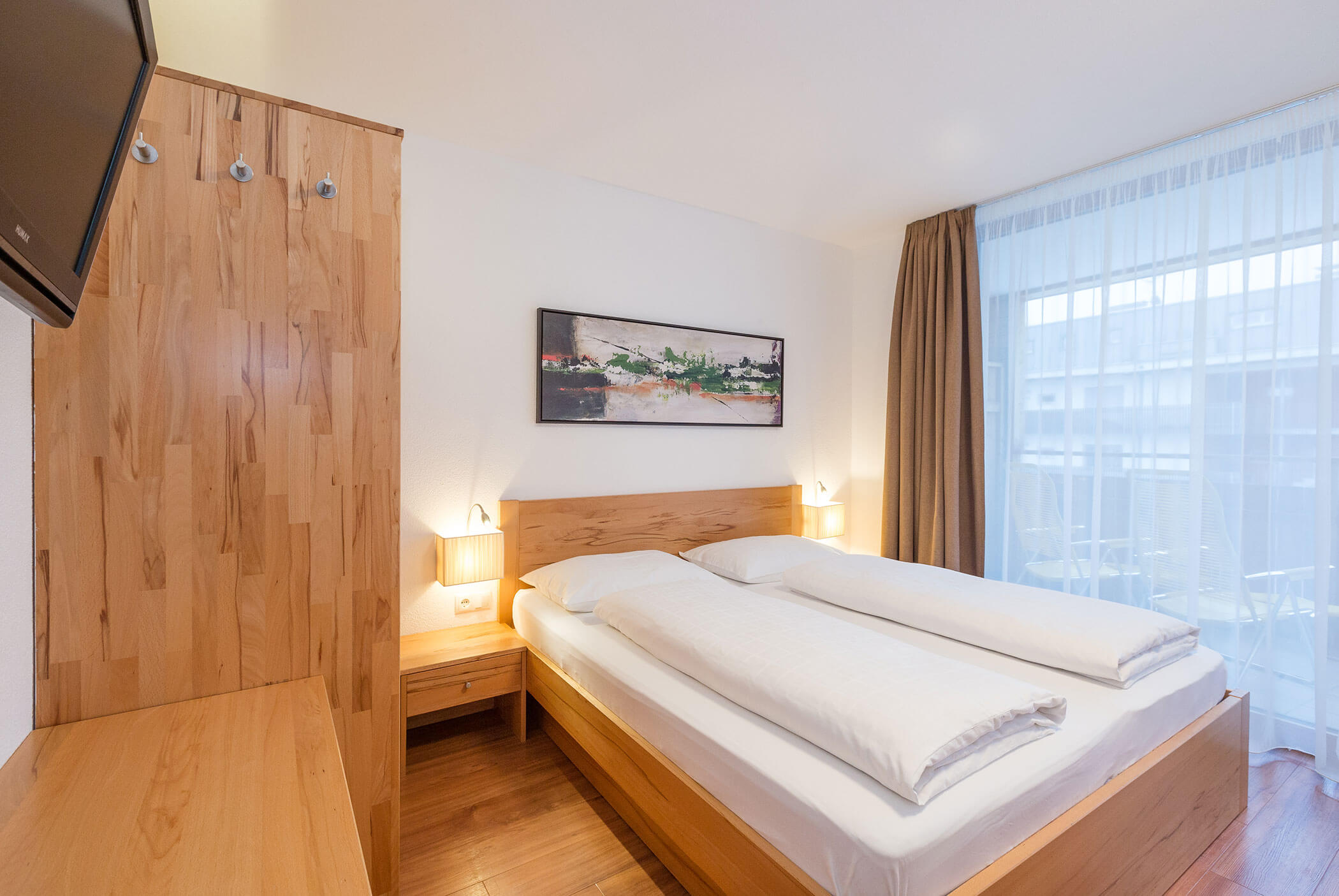 Camera d'albergo luminosa con letto matrimoniale, mobili in legno, quadri alle pareti e un'ampia finestra con tende trasparenti. - Residence Aichner