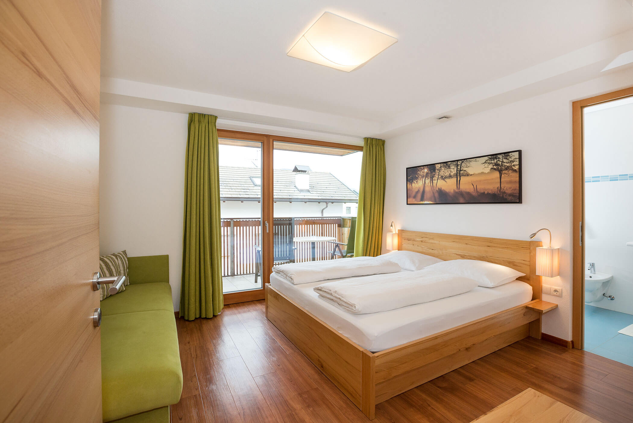 Camera da letto luminosa con mobili in legno, tende verdi, balcone e un bagno visibile sulla destra. - Residence Aichner