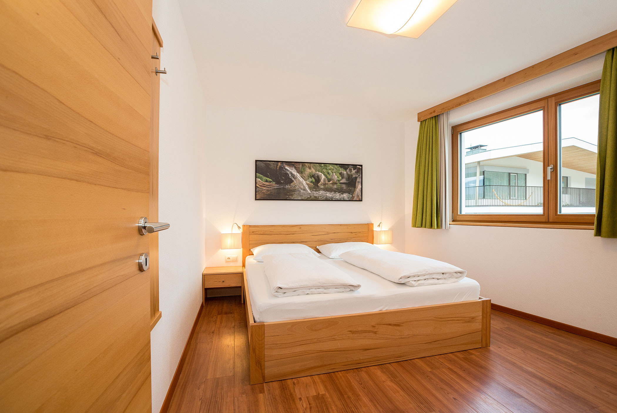 Camera da letto in stile minimalista con letto in legno, due comodini, un'ampia finestra e un quadro raffigurante la natura sopra il letto. - Residence Aichner